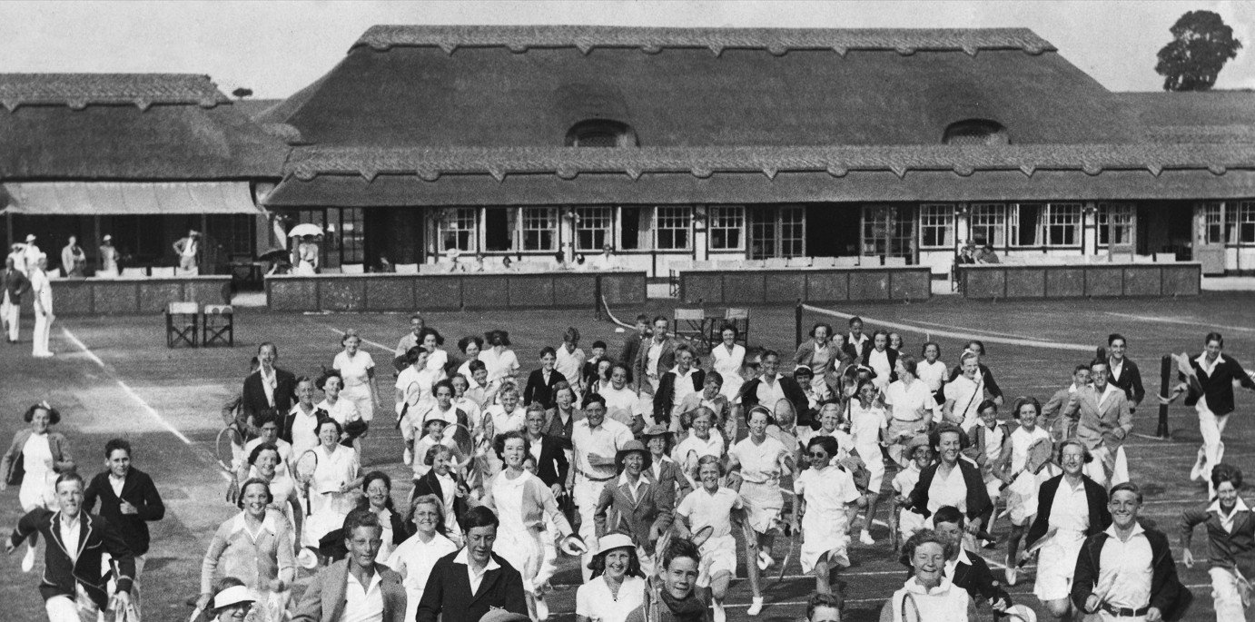 Black and white photo of a large group of women and girls running on a grassy field, likely at a sports event, in front of a large building with a thatched roof.