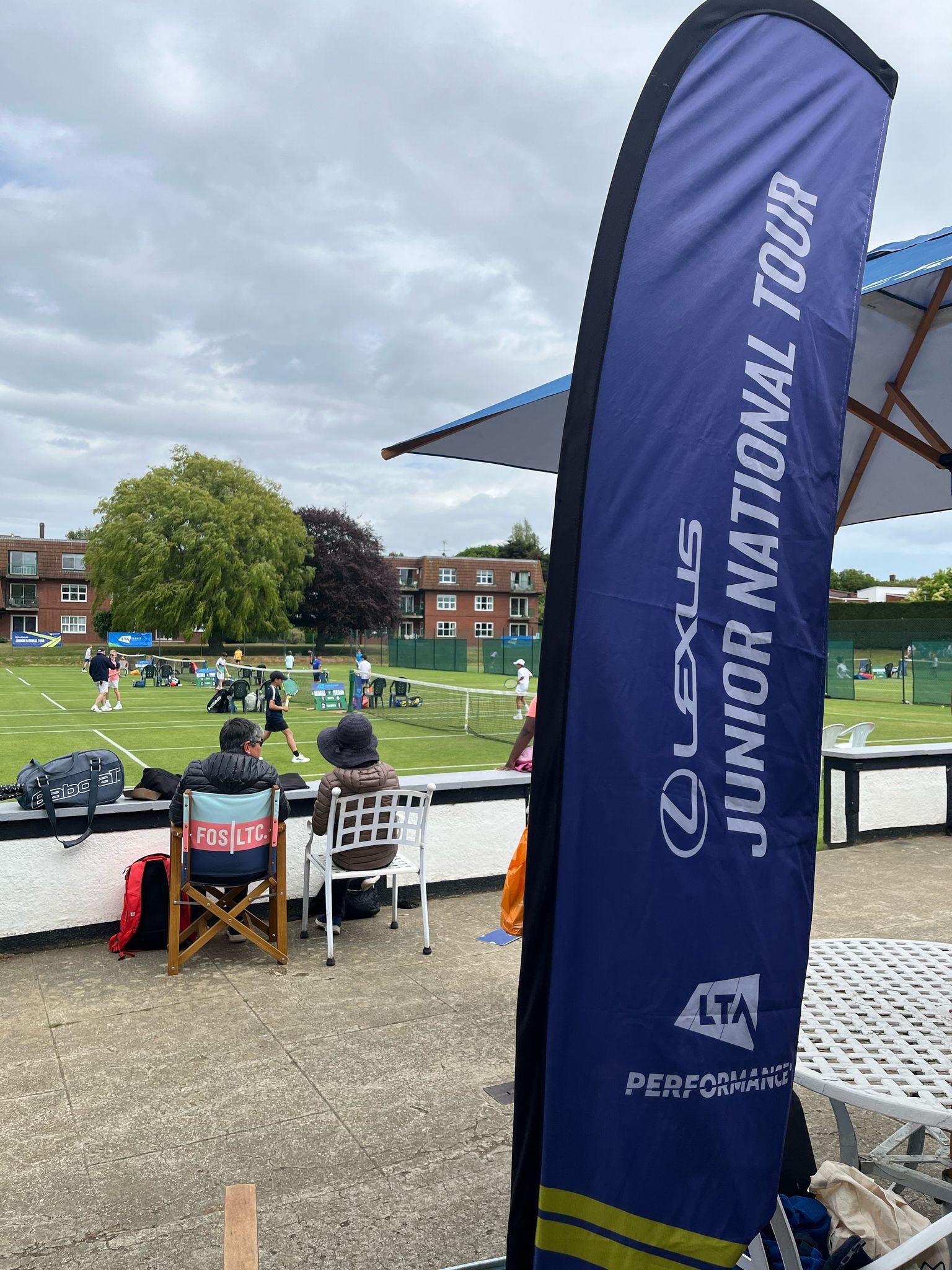 People playing tennis on a clay court with some spectators sitting nearby, a large blue banner with 'UNION NATIONAL TOUR' and 'LTA PERFORMANCE' written on it, and a residential building in the background.