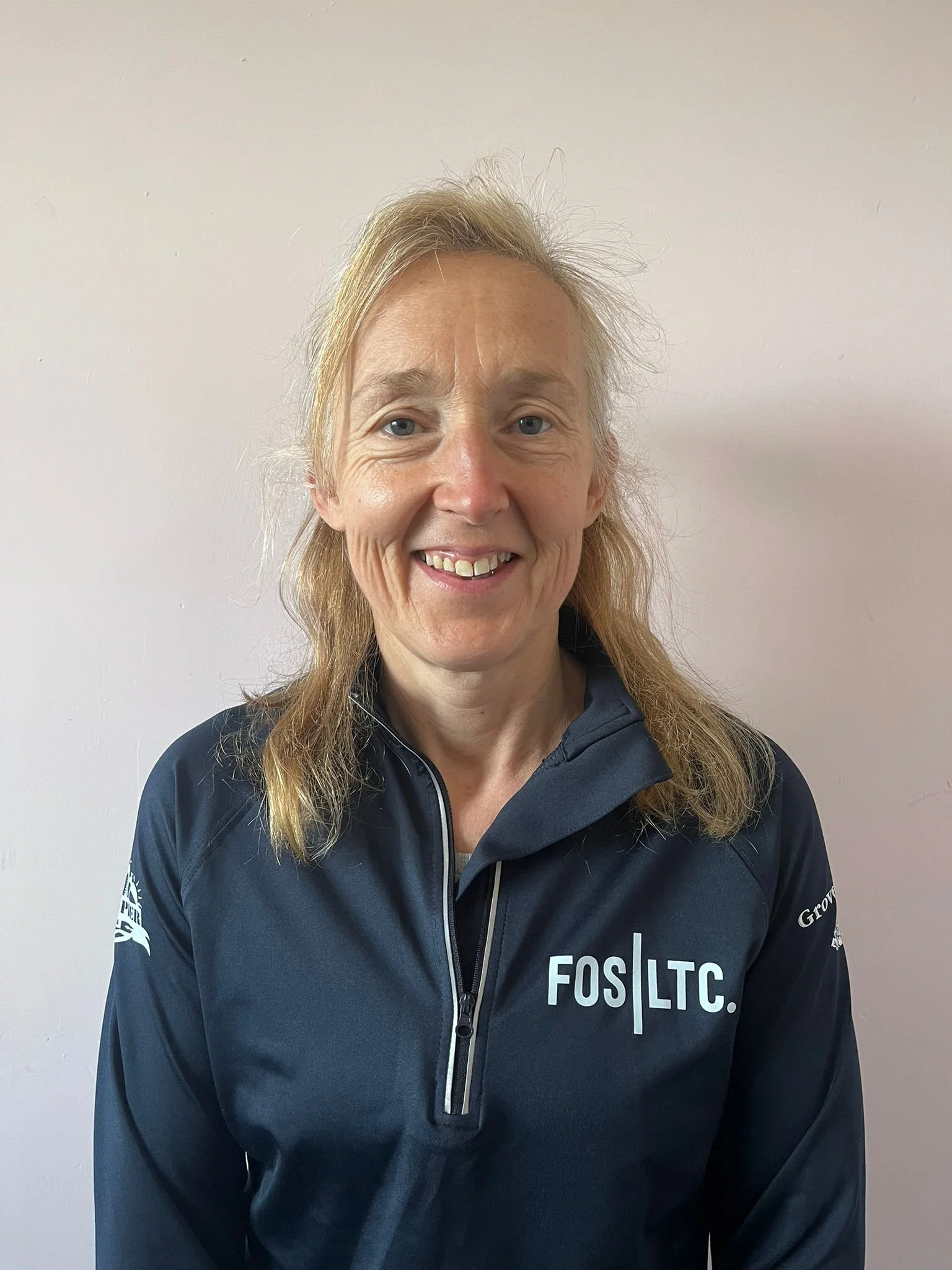 A smiling woman with blonde hair wearing a navy blue jacket with a logo that says 'FOS|LTC' on the front, standing against a plain light-colored wall.