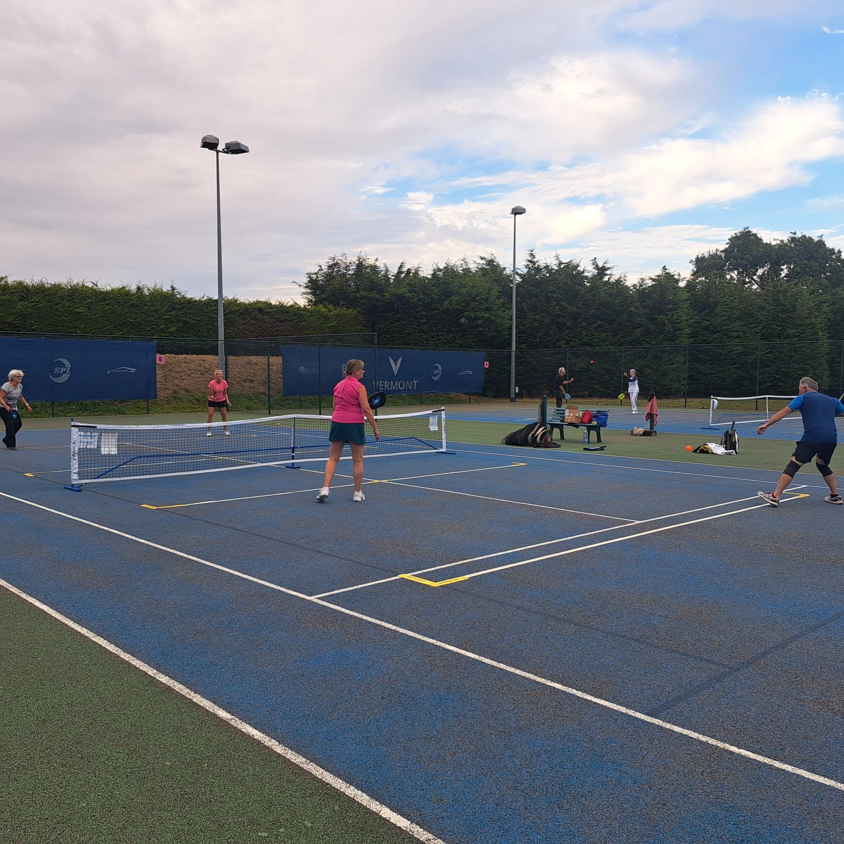People playing pickleball on an outdoor court surrounded by trees and a cloudy sky.