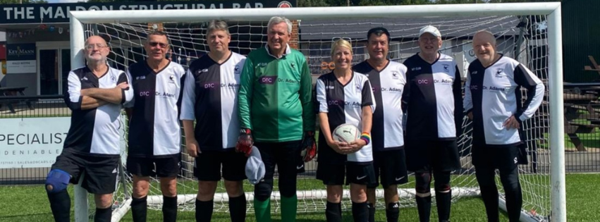 Group of eight people standing in front of a soccer goal on a field, dressed in soccer uniforms, one wearing goalkeeper gear and the others in matching team jerseys, some holding a soccer ball.