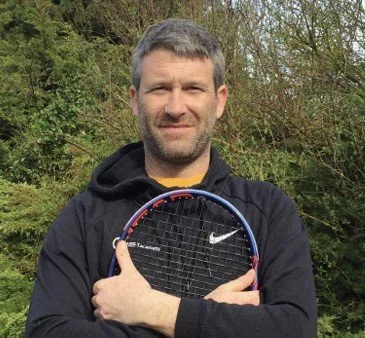 A man with short, gray hair and a beard, wearing a black Nike hoodie, holding a tennis racket in front of him with both arms crossed, outdoors in a natural setting.