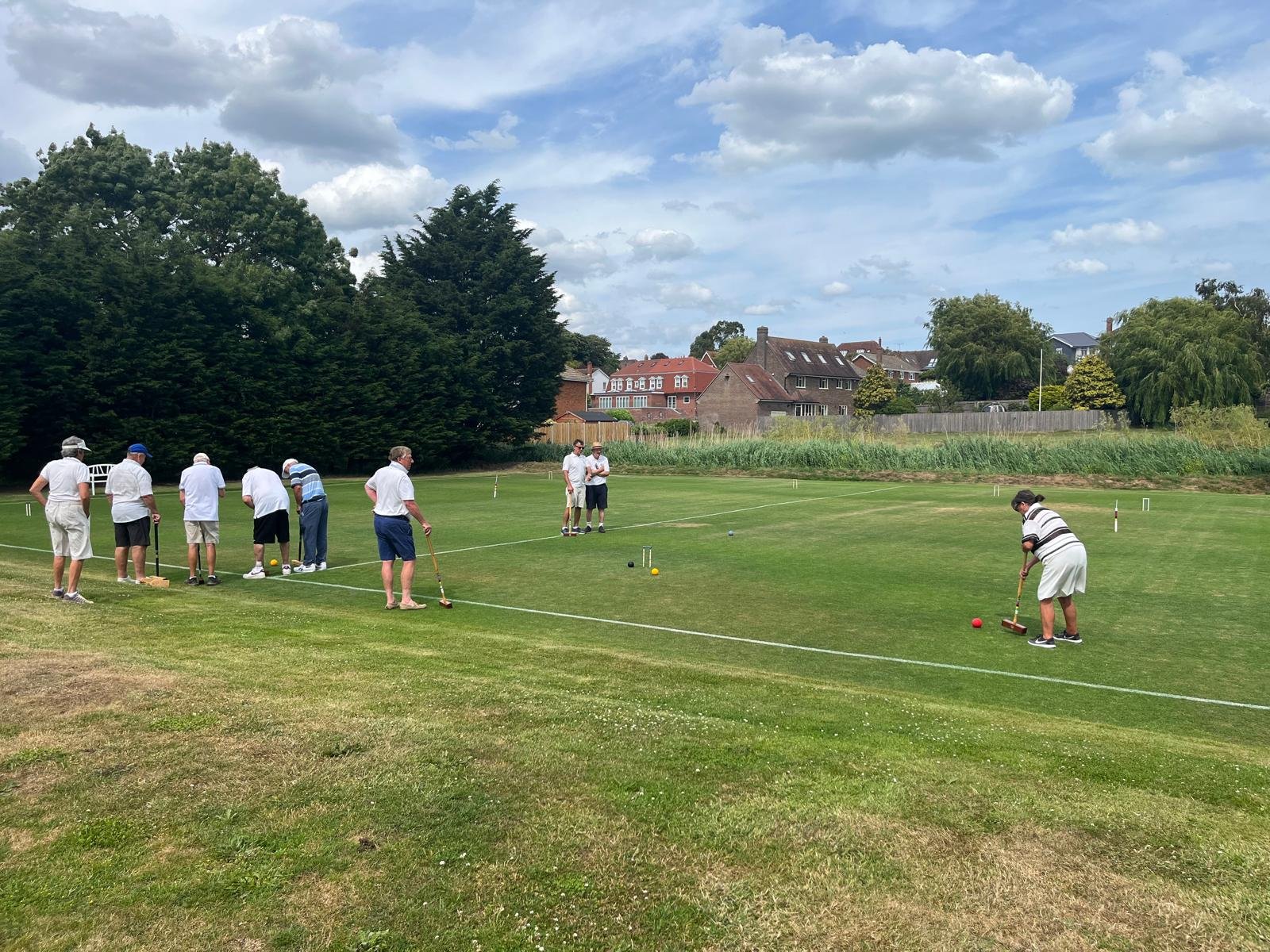 Group of people playing croquet on a grassy field with trees and houses in the background under a partly cloudy sky.