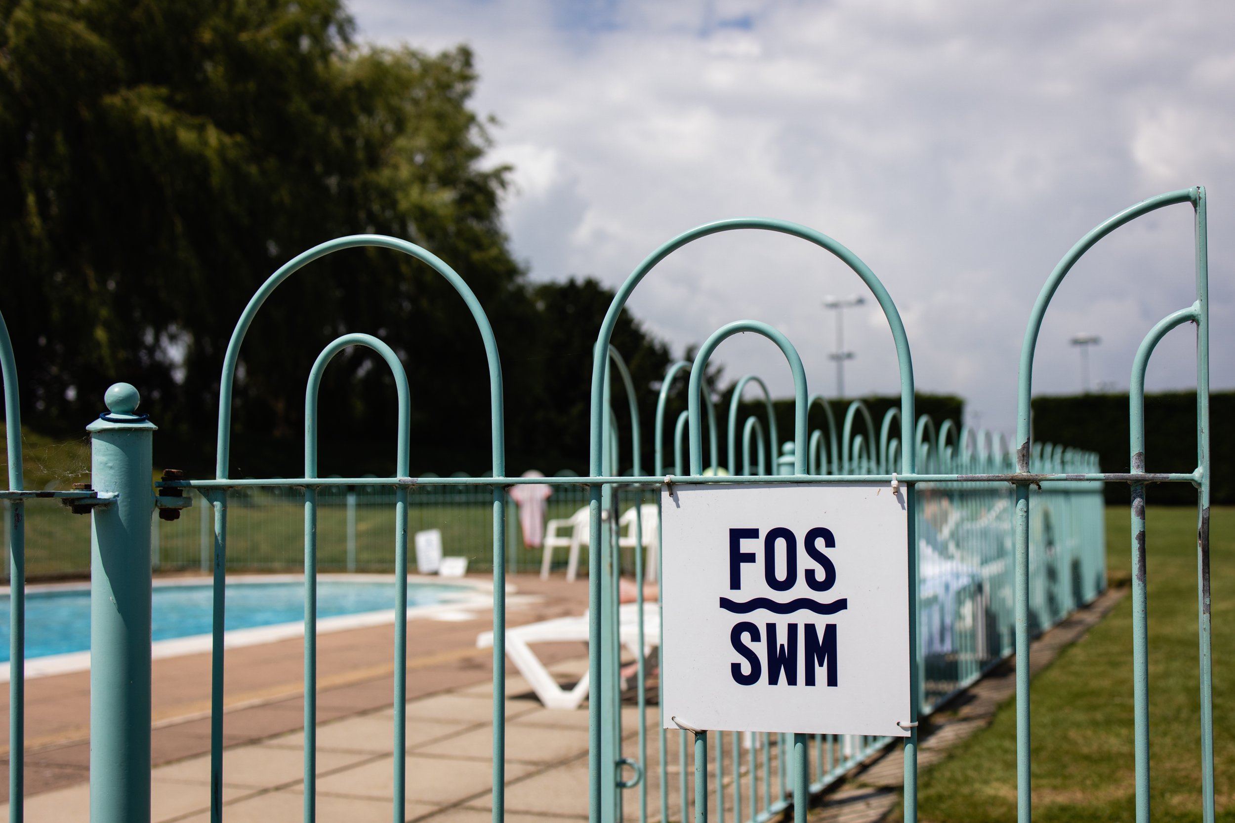 A swimming pool area enclosed by a light blue metal fence with a sign that reads 'FOS SWIM'. The pool has a wooden deck with pool chairs, and the background includes trees, grass, and a cloudy sky.