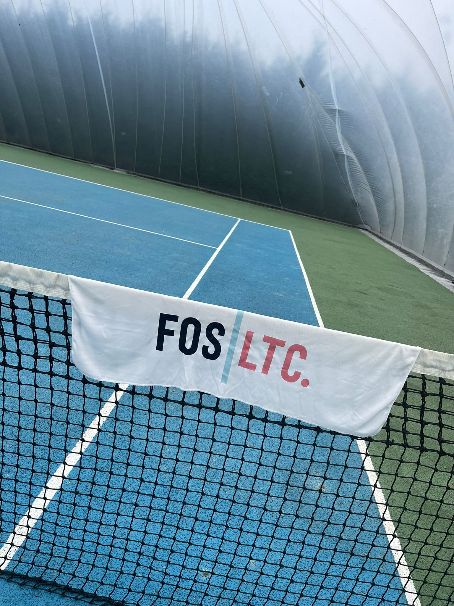 Indoor tennis court with a blue surface and a black net in the foreground, with a banner hanging on the net reading 'FOS | T.C.'