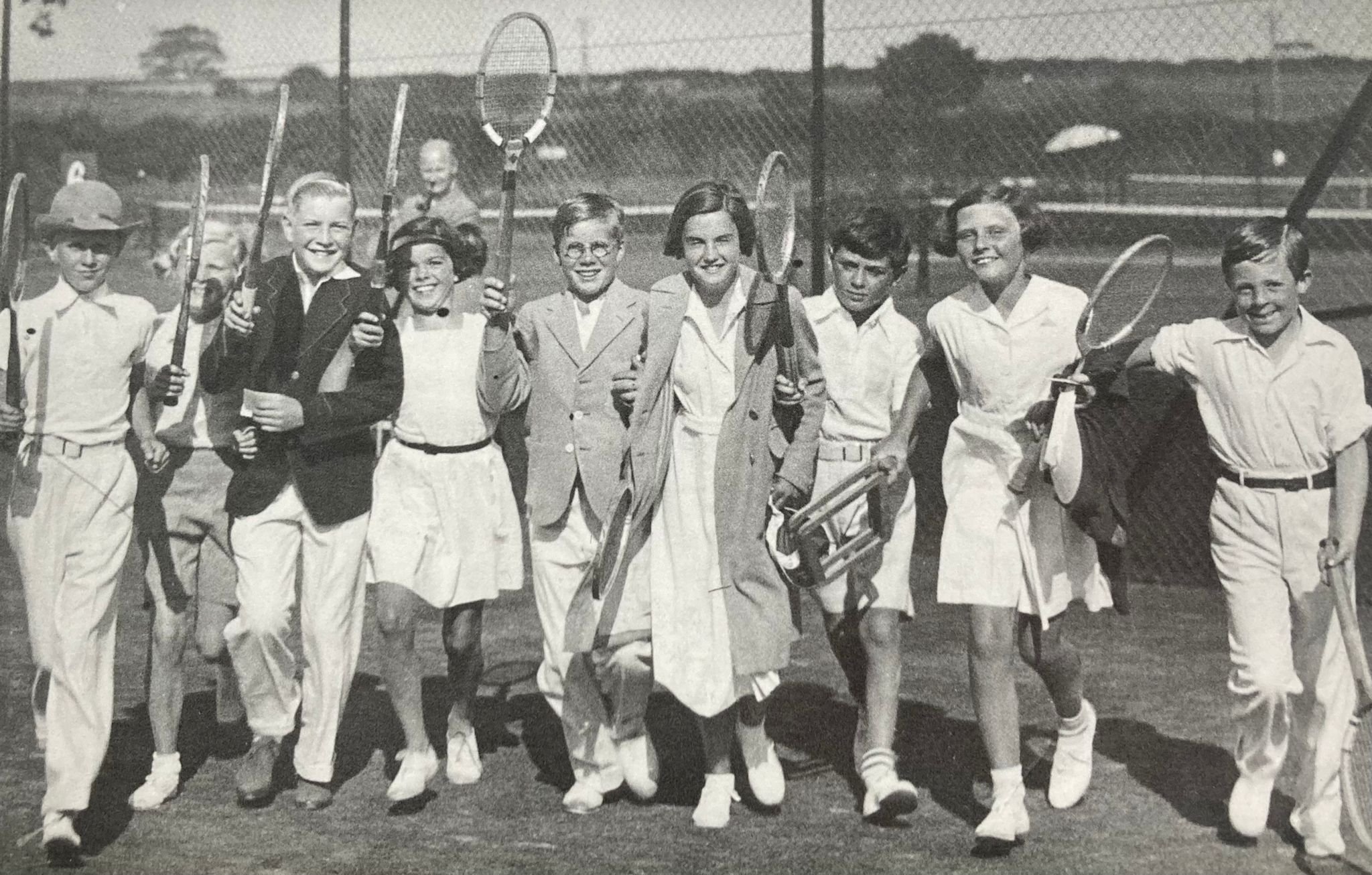 Black-and-white photo of children at a tennis court, smiling and holding tennis rackets, dressed in vintage clothing.