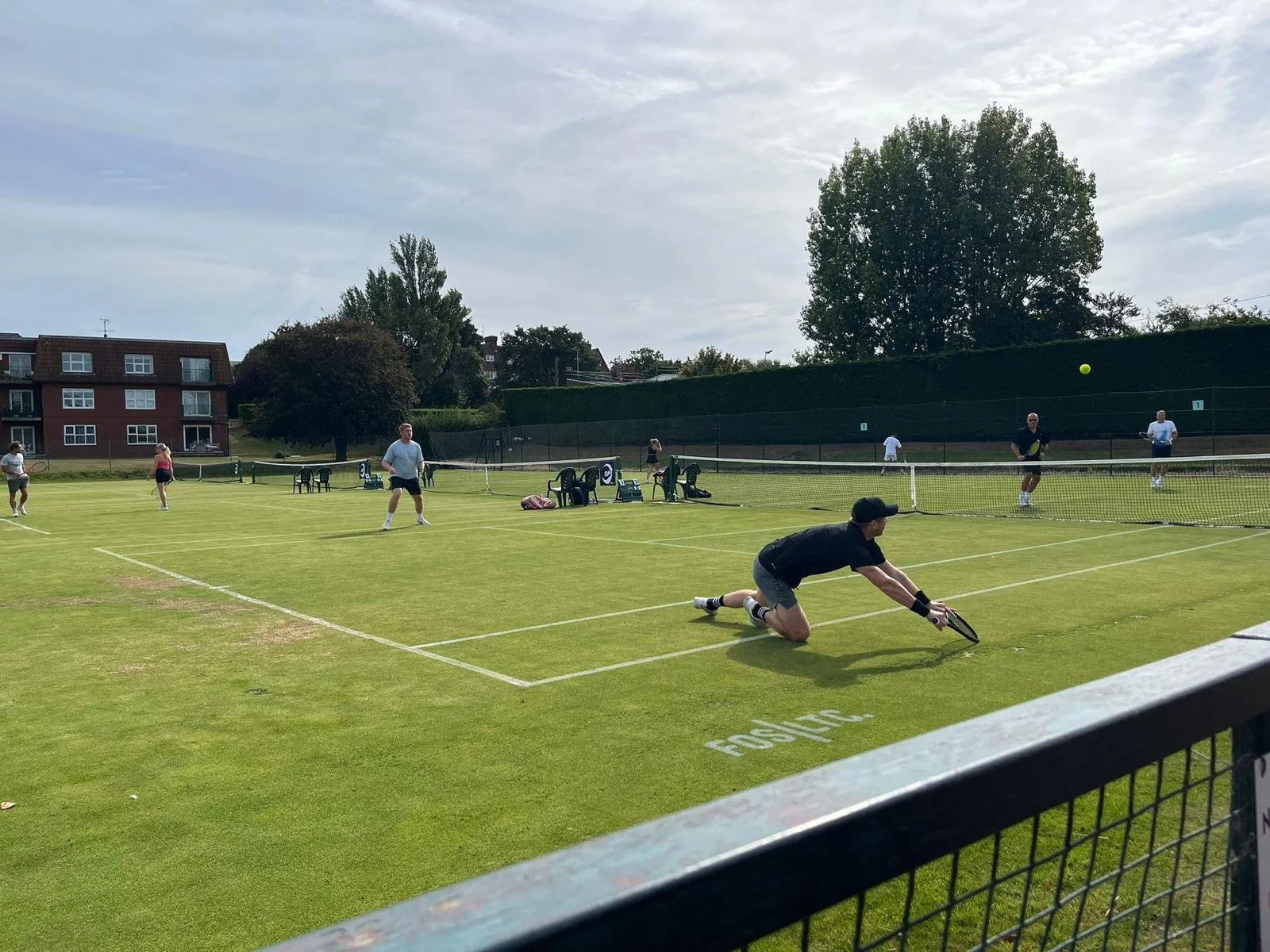 A group of people playing tennis on a grass court on a sunny day, with multiple players preparing to hit the ball and a man in black diving to reach the ball.