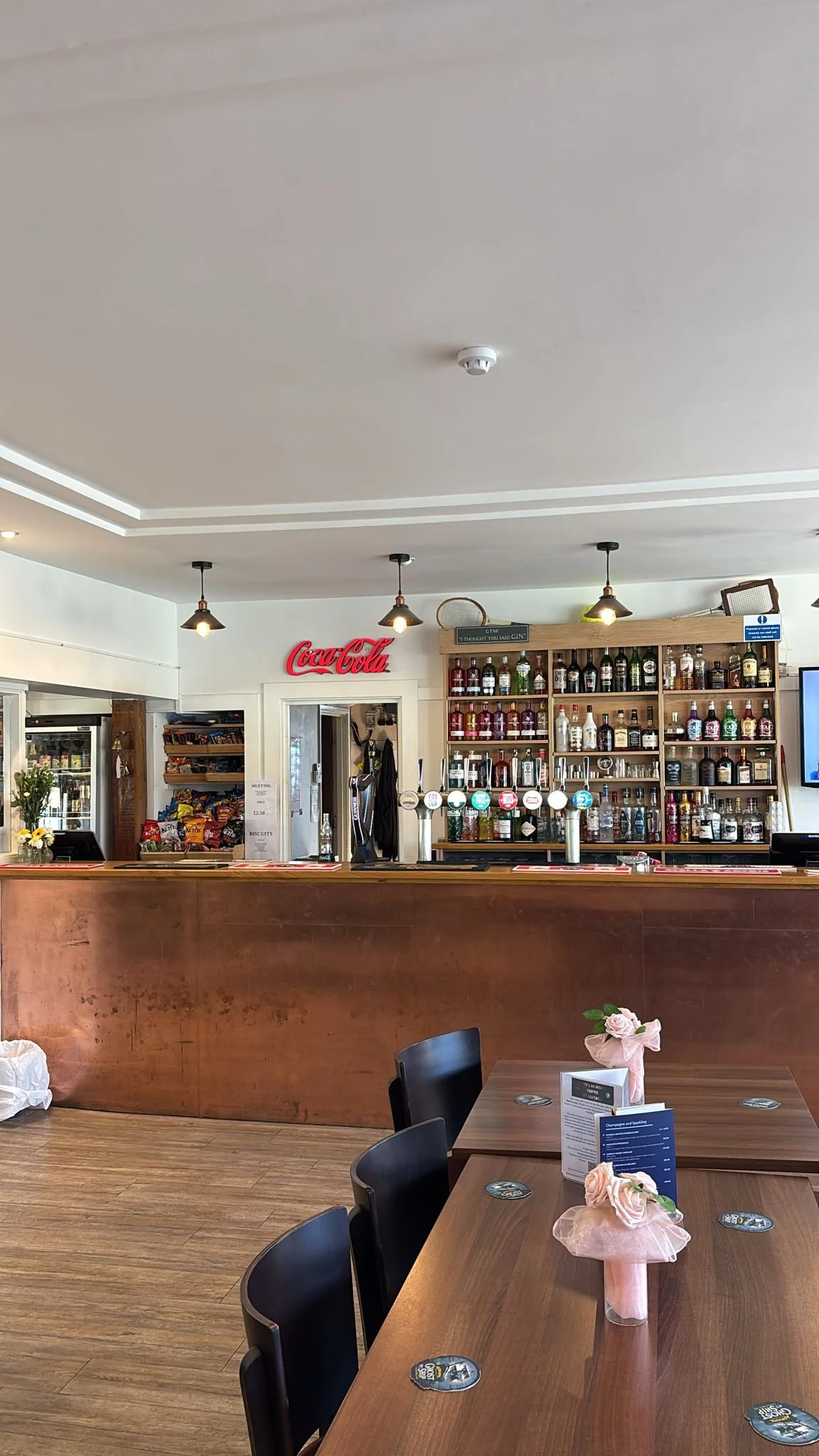 Interior of a bar or restaurant with a wooden counter, shelves with bottles of alcohol, a Coca-Cola sign, tables with pink flower arrangements, and a TV screen.