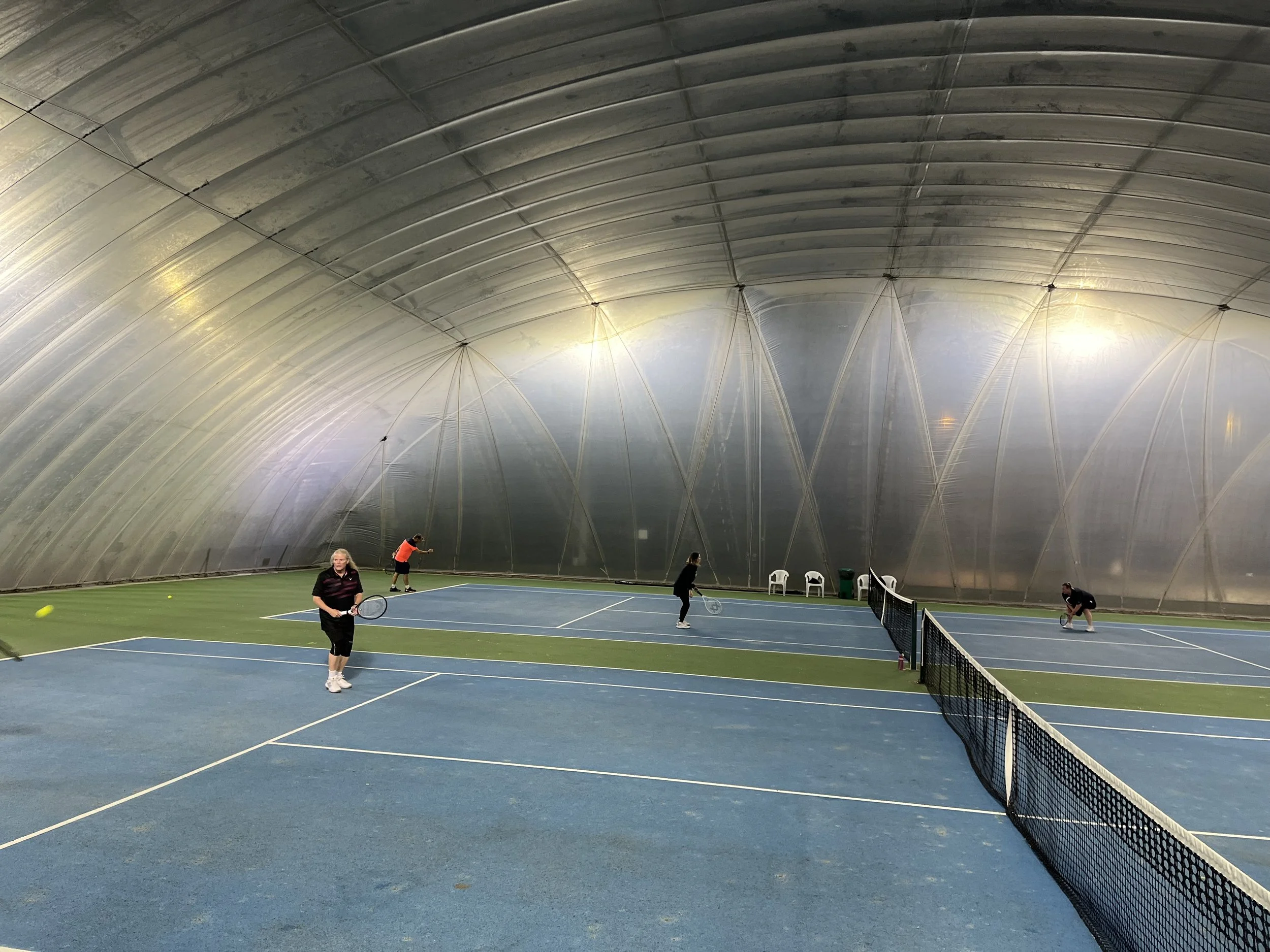 Indoor tennis court with four people playing tennis, two near the net and two at the back of the court, under a high, curved ceiling.