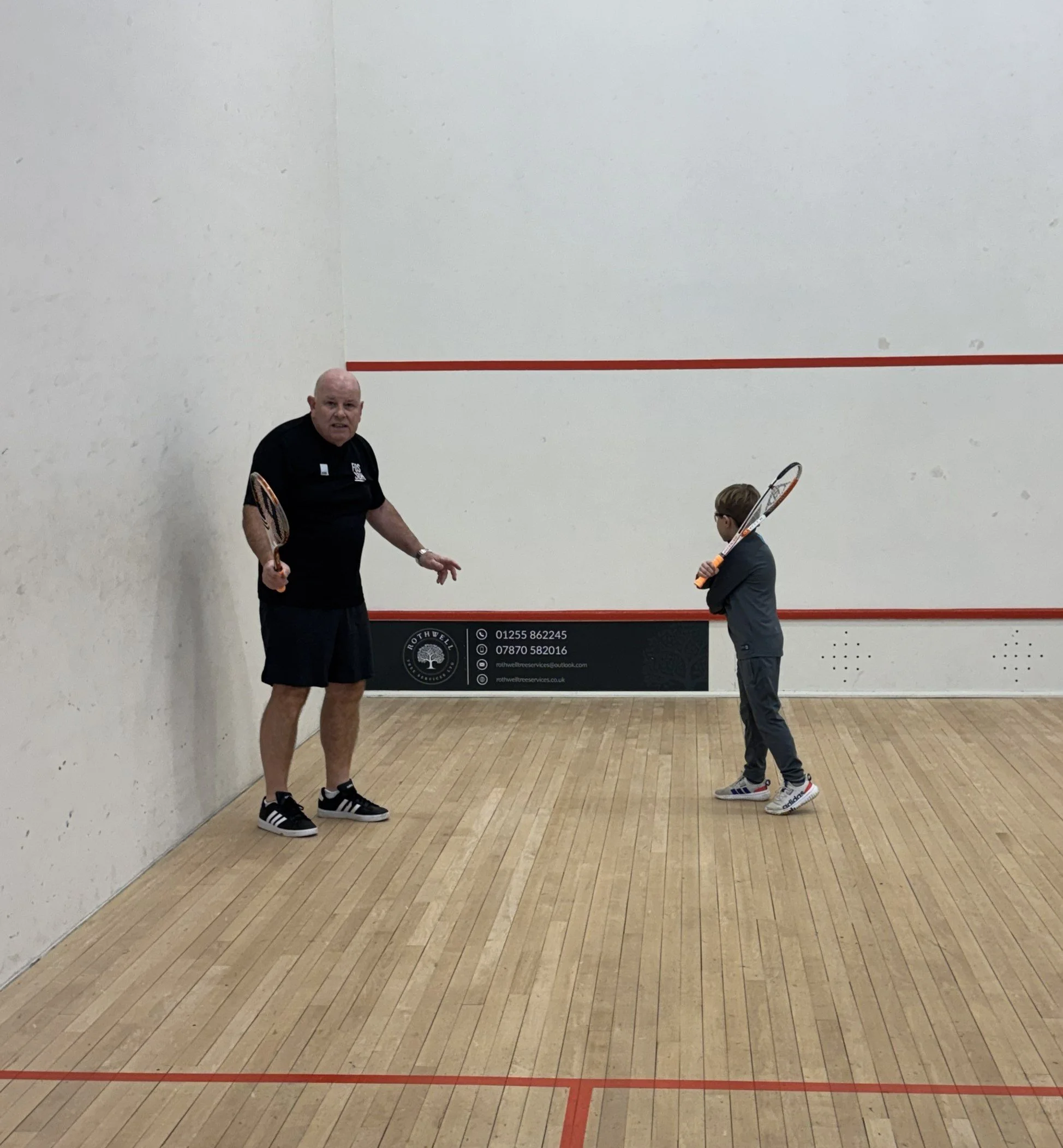 An adult and a child playing squash on an indoor court. The adult is holding a squash racket and appears to be instructing or coaching, while the child is also holding a racket and facing the adult.