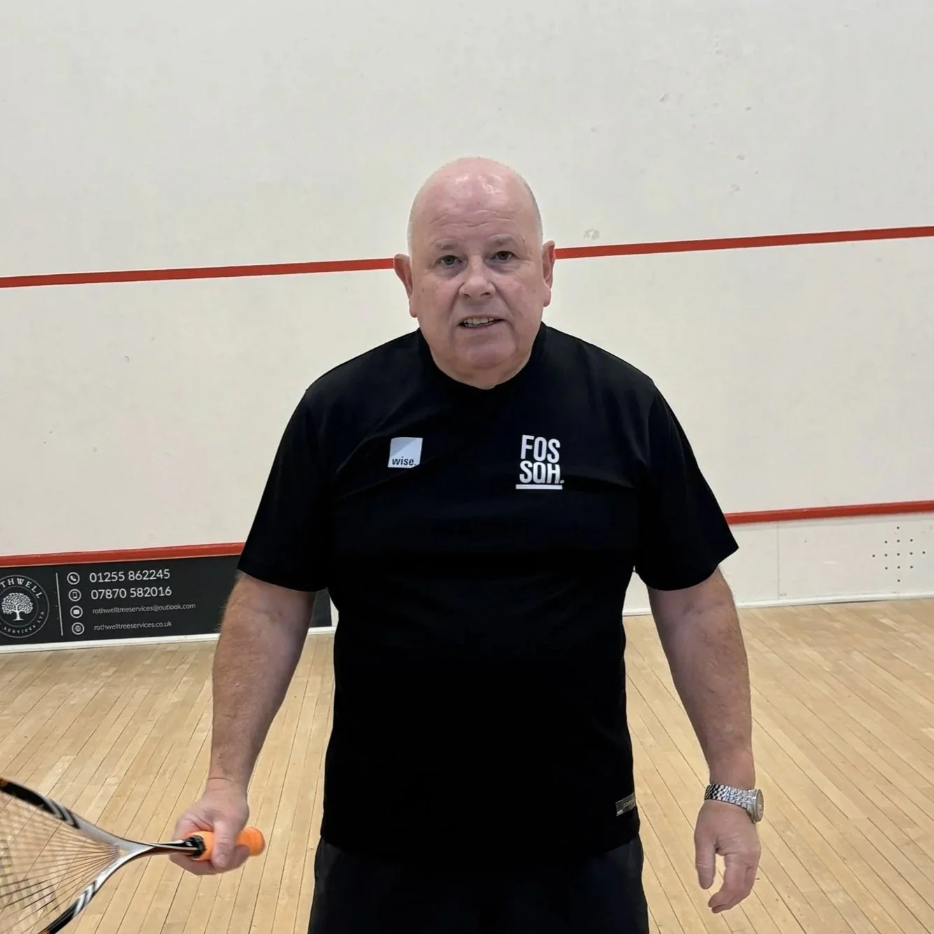 An elderly man playing squash, holding a squash racket, inside a squash court.