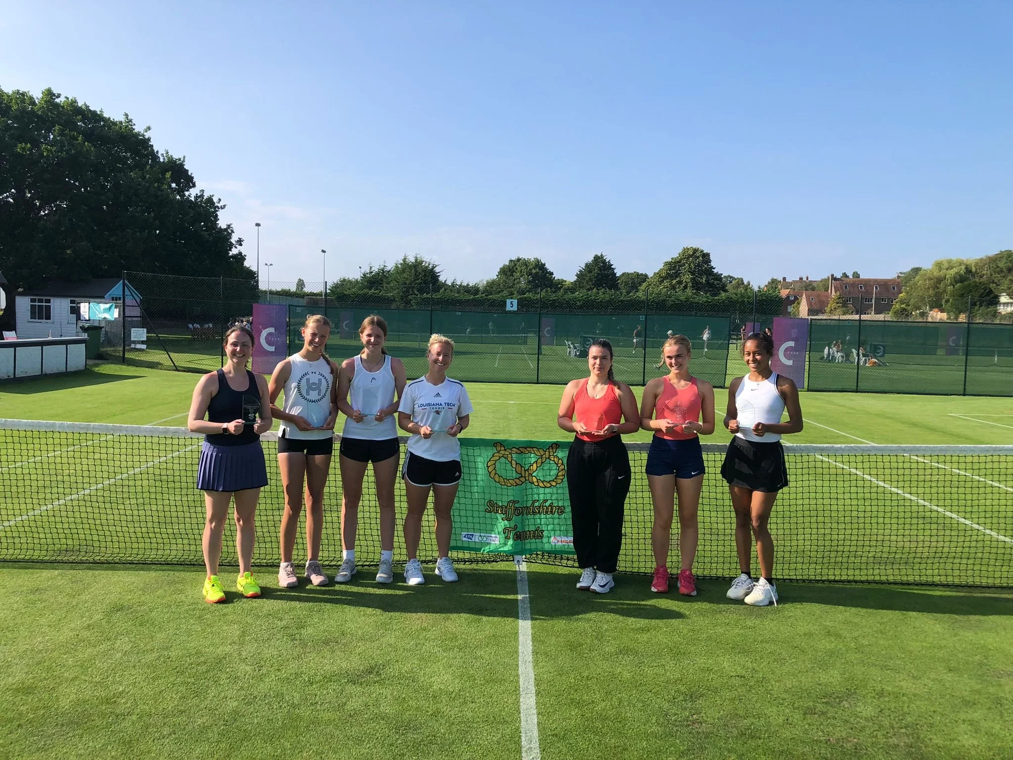 Group of eight female tennis players standing behind a net on a grass tennis court, holding awards, with trees and residential houses in the background on a sunny day.