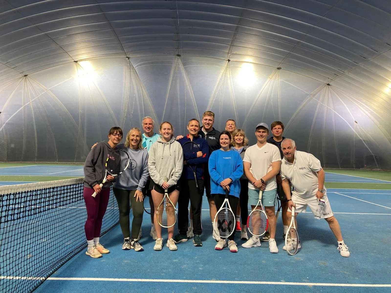 Group of people on an indoor tennis court holding tennis rackets, under a large domed ceiling.