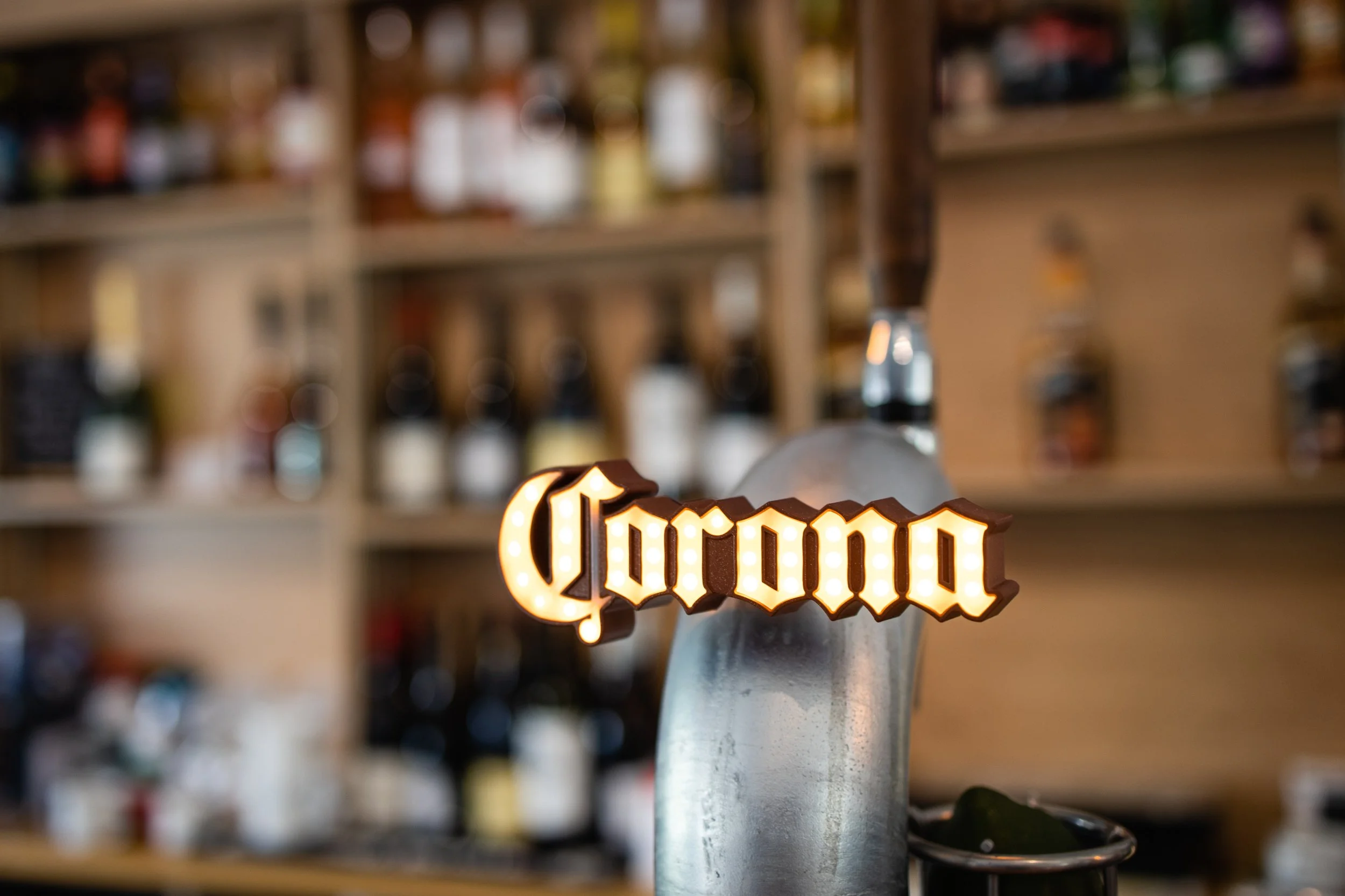 Close-up of a Corona beer tap handle with the illuminated "Corona" logo on it, in a bar setting with shelves of liquor bottles in the blurred background.