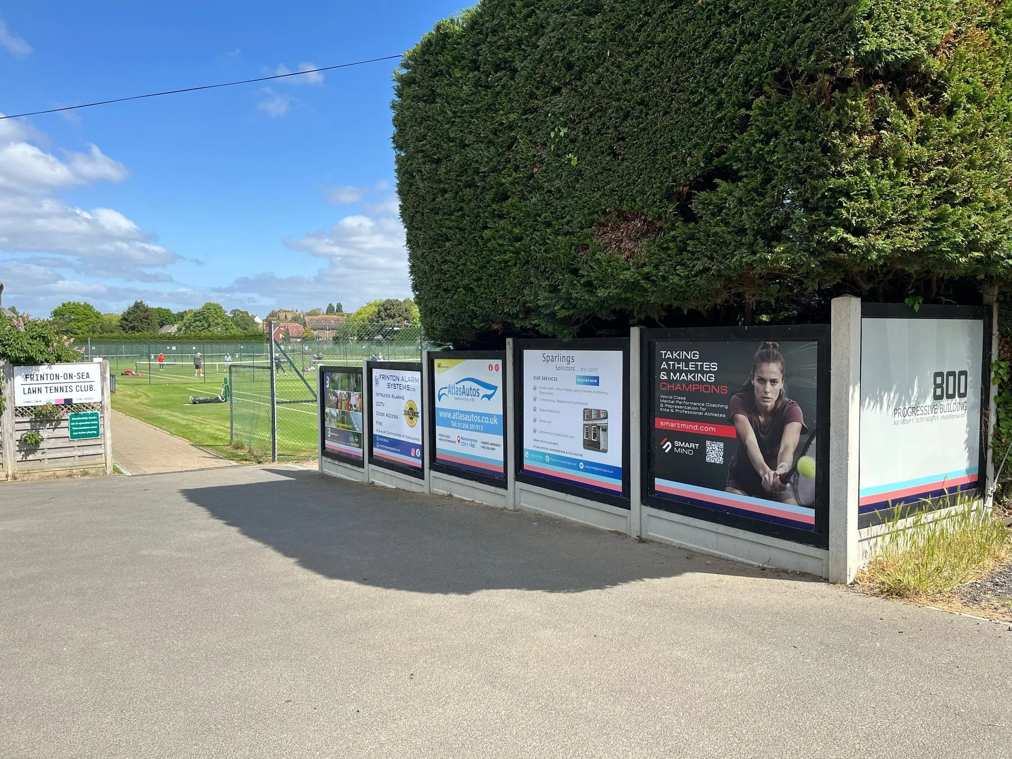 View of a sports field with people playing tennis, fencing around the field, signboards on the gate, and advertisements on the perimeter, with a clear blue sky and a large green tree.