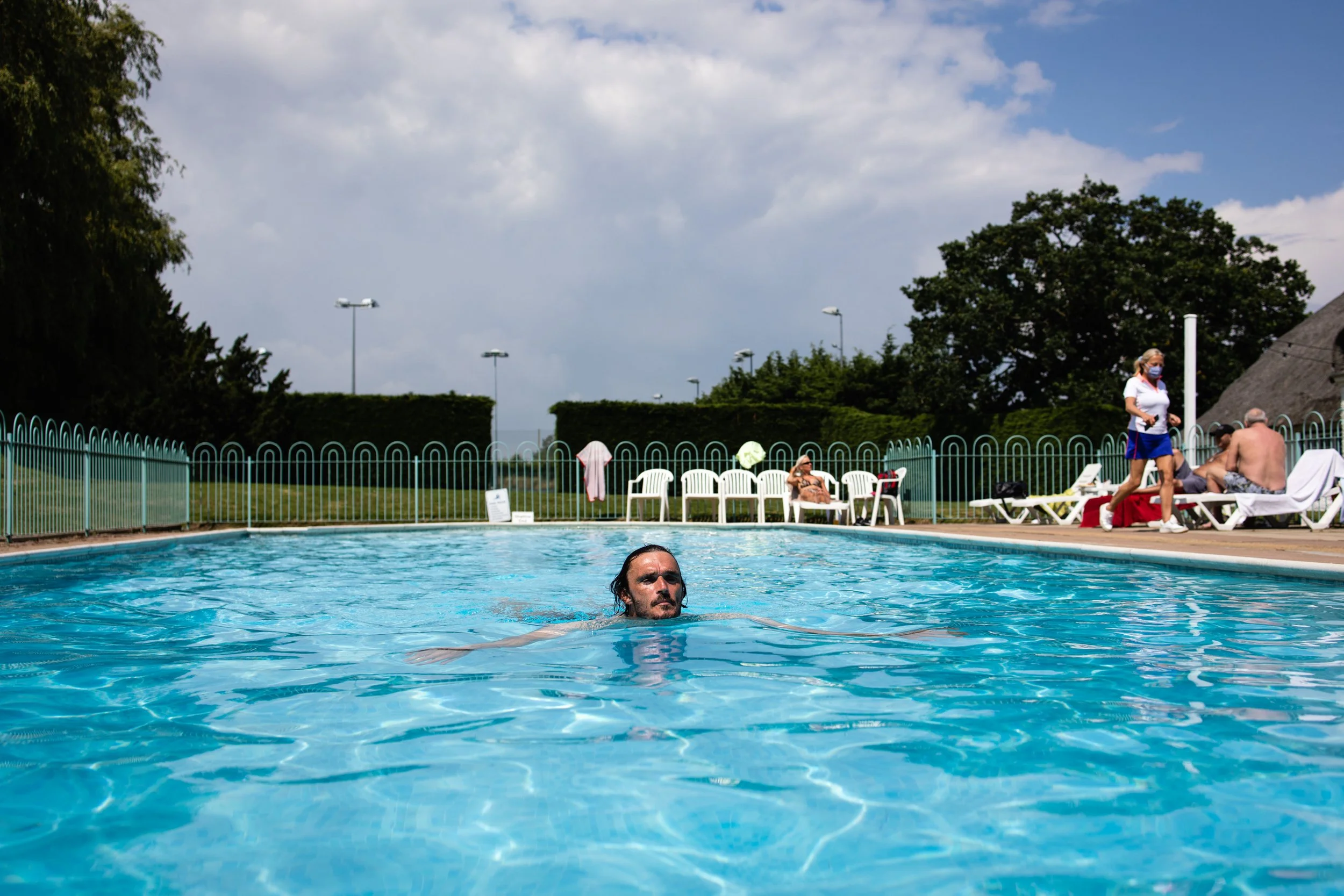 Man swimming in a blue outdoor pool on a sunny day, with people lounging on chairs and umbrellas in the background.