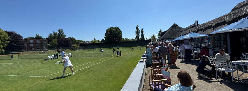 People watching a lawn bowling game on a sunny day at an outdoor venue with a building and trees in the background.