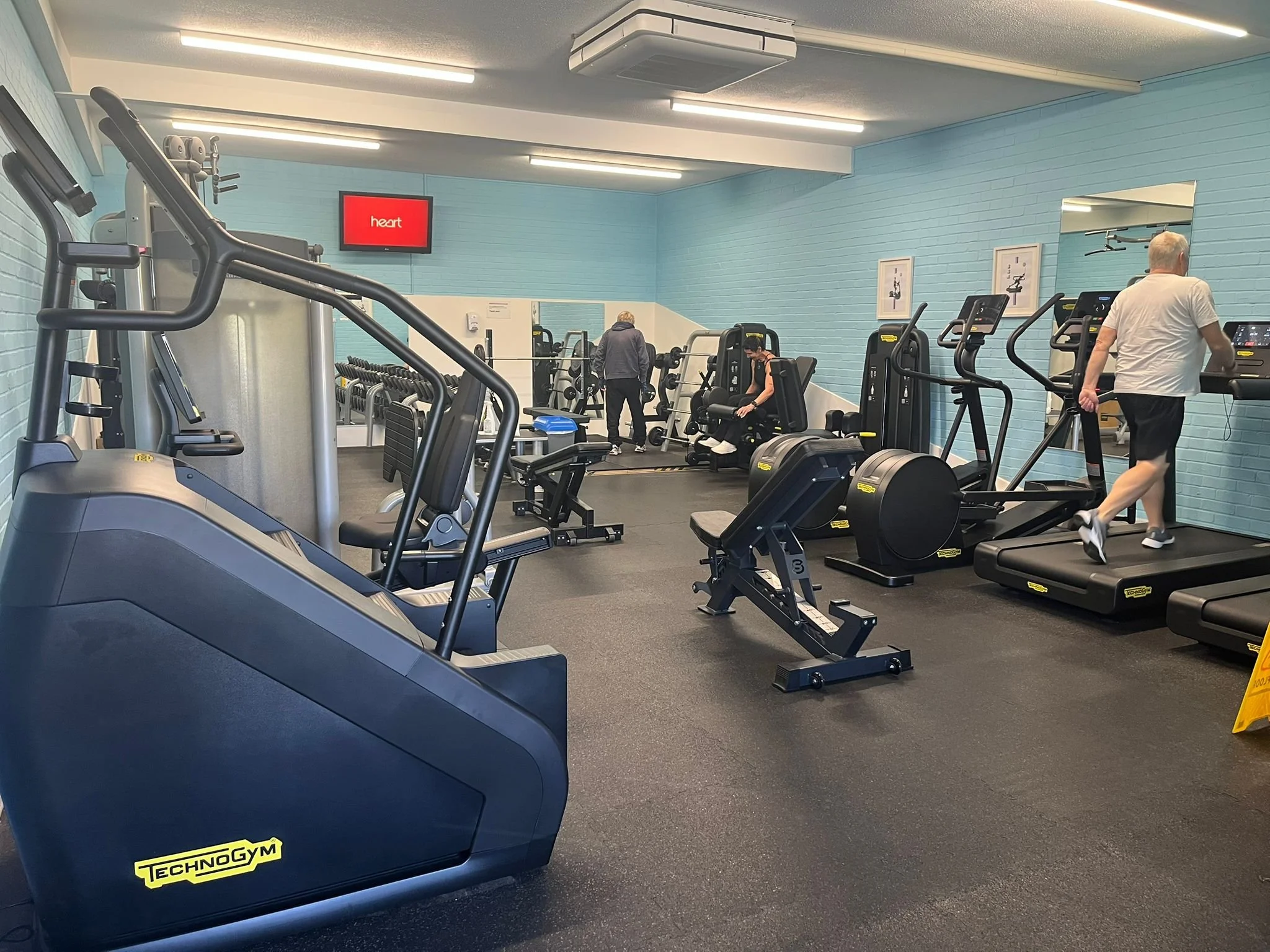A gym with various exercise equipment including treadmills, ellipticals, and strength machines. There are two people working out and a woman sitting on a seated chest press machine. The walls are painted light blue, and there is a mirror on one wall with a red TV screen above it showing the word "heart."