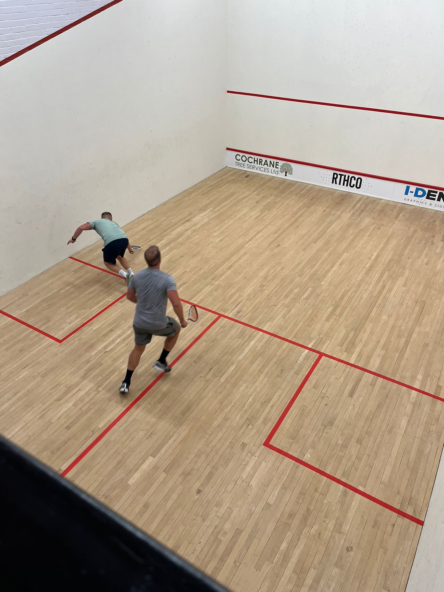 Two men playing indoor racquetball on a wooden court with red boundary lines. One man is near the back wall, reaching for the ball, while the other is closer to the camera, holding a racquet.