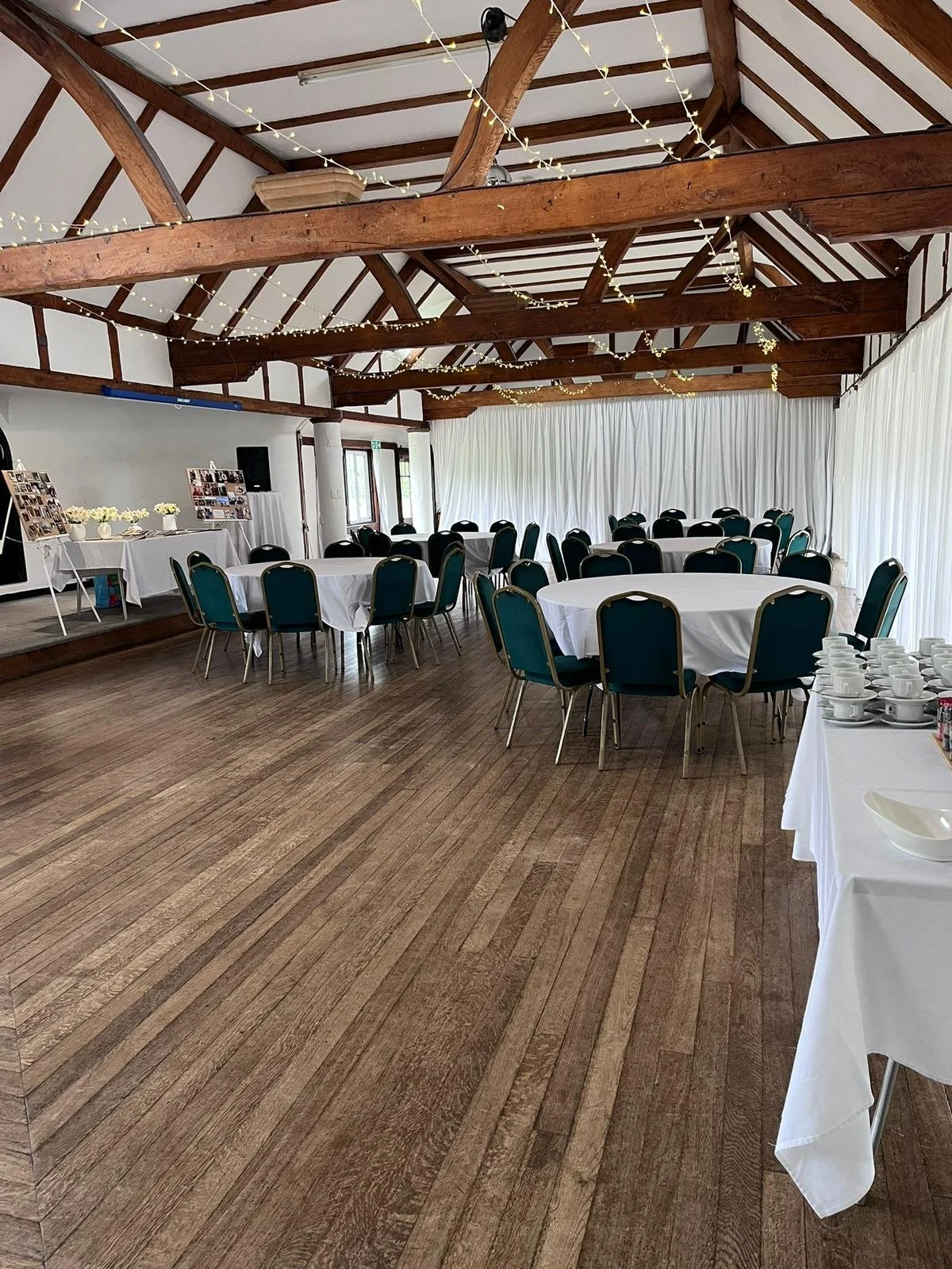 Event space with round tables covered in white tablecloths and teal chairs in a wooden hall, decorated with string lights and set up for a celebration.