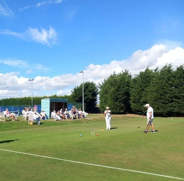People playing and watching lawn bowls on a well-maintained green with a blue sky and trees in the background.
