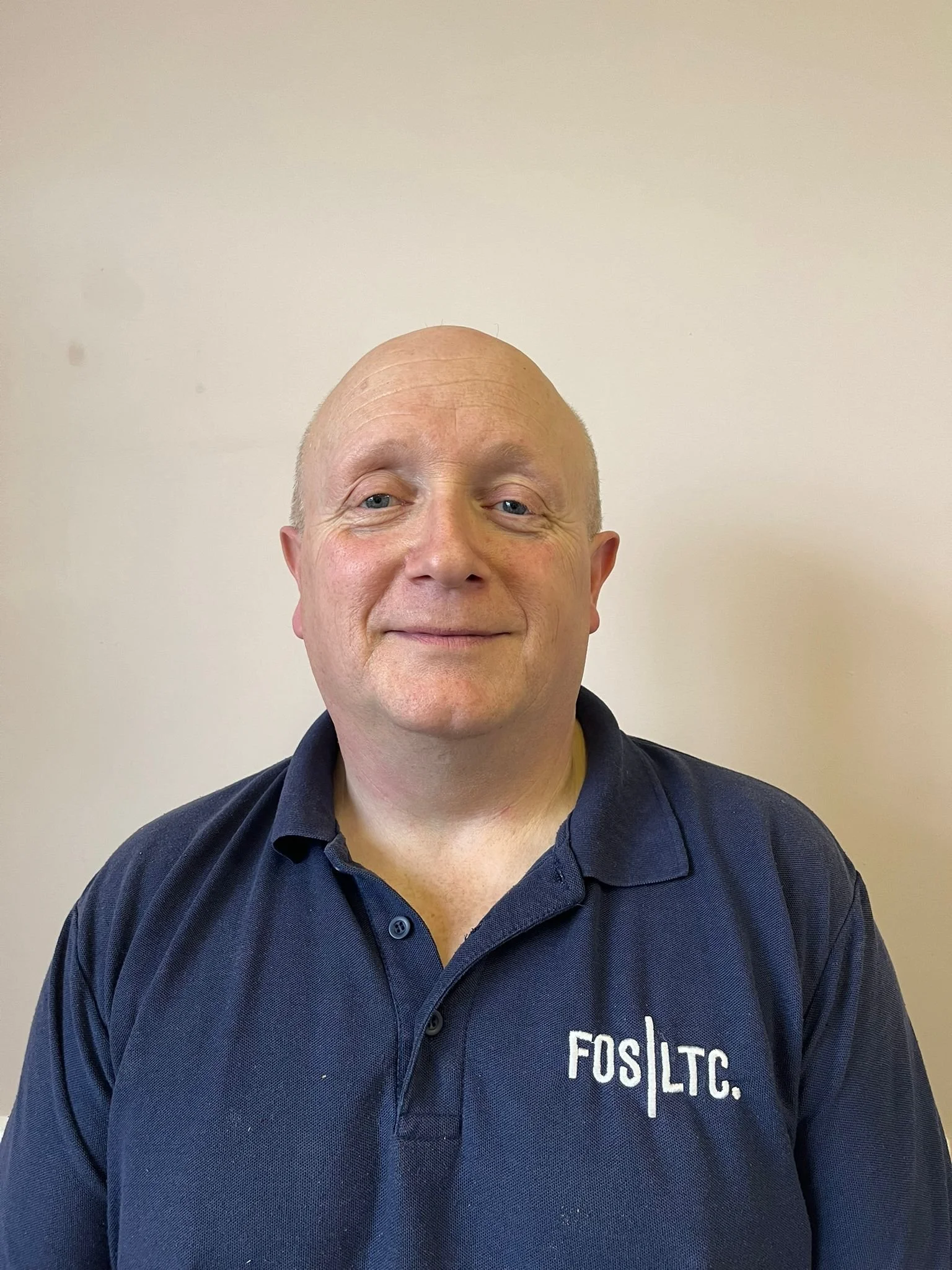 A middle-aged man with a bald head and blue eyes smiling, wearing a navy blue polo shirt with 'FOS LTC.' embroidered on the chest, standing against a plain light-colored wall.