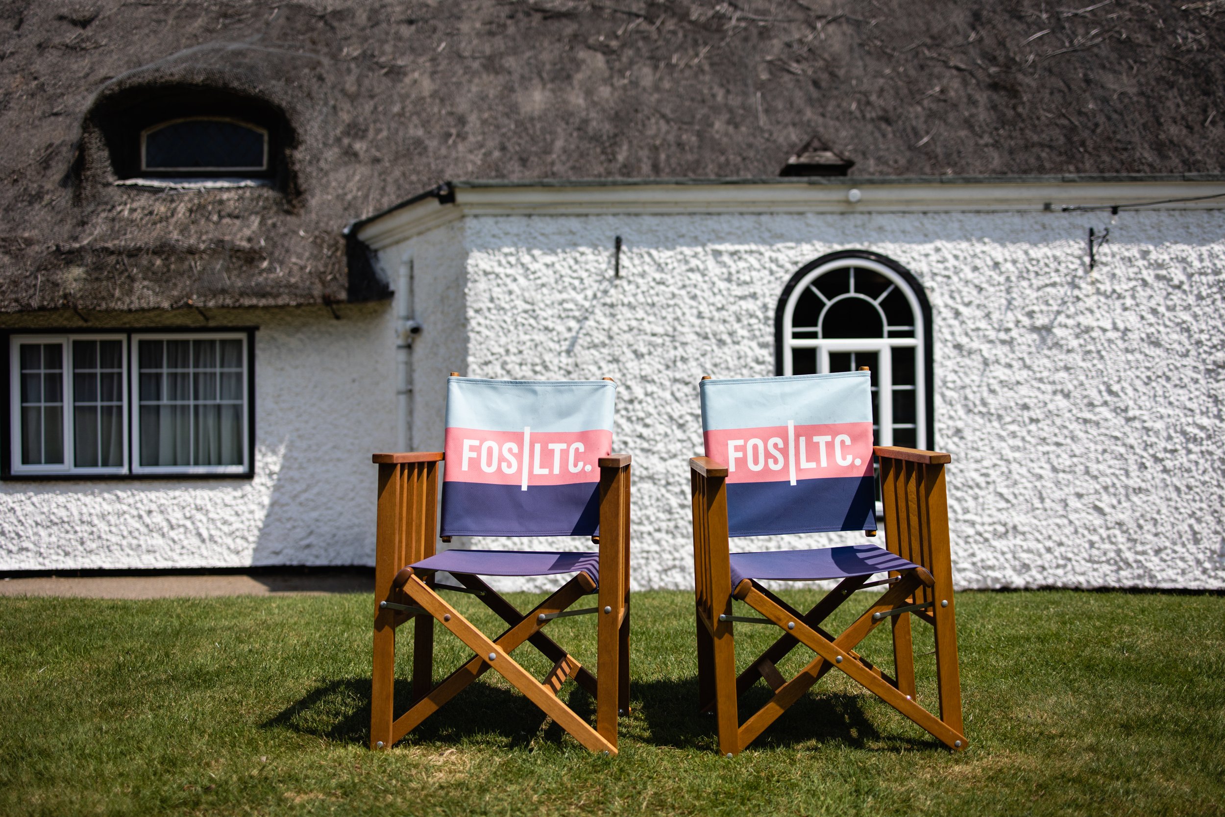 Two outdoor chairs with wooden frames and fabric seats, both displaying the logo 'FOS LTC' in pink and white, placed on a grassy lawn in front of a white textured house with a thatched roof and arched windows.