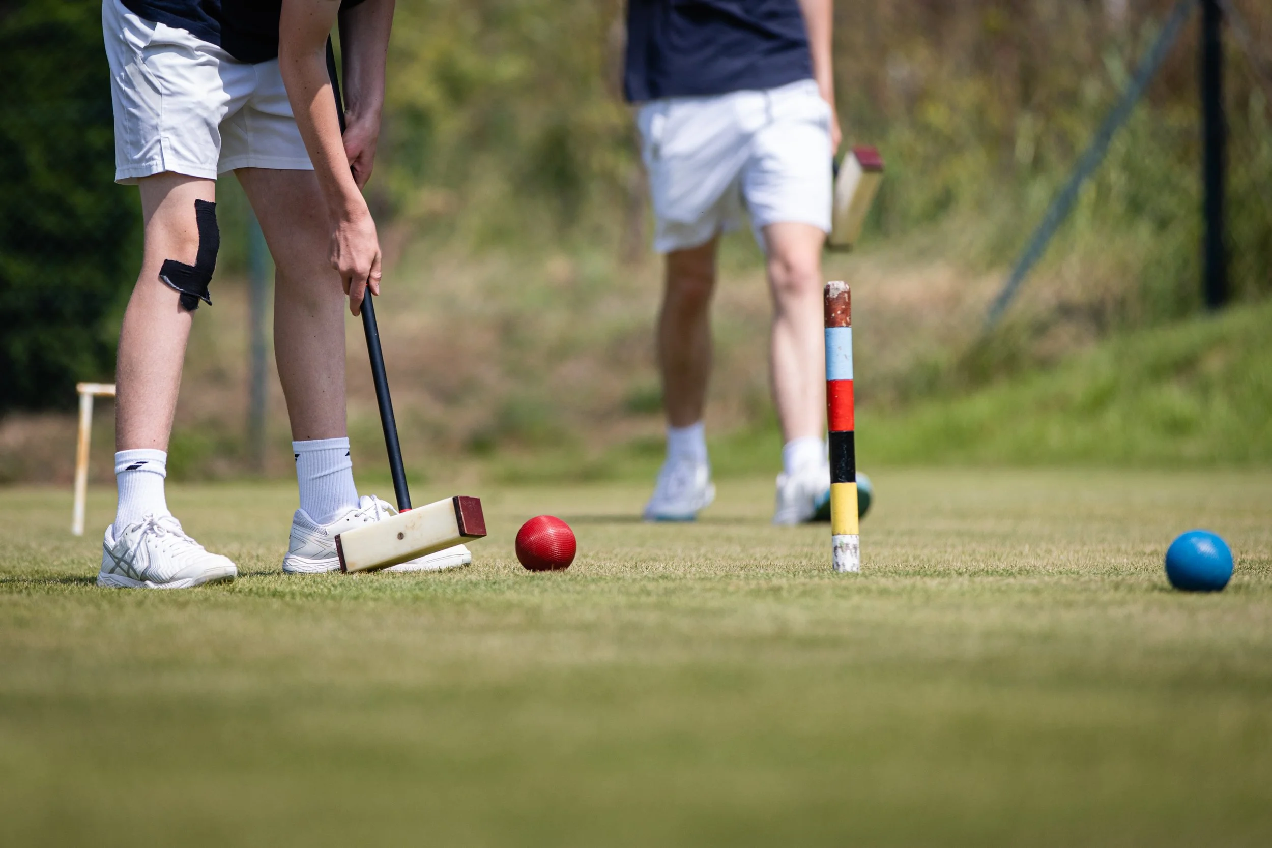 Two people playing croquet on a grass field, with one person preparing to hit a red ball using a mallet, while the other stands nearby.