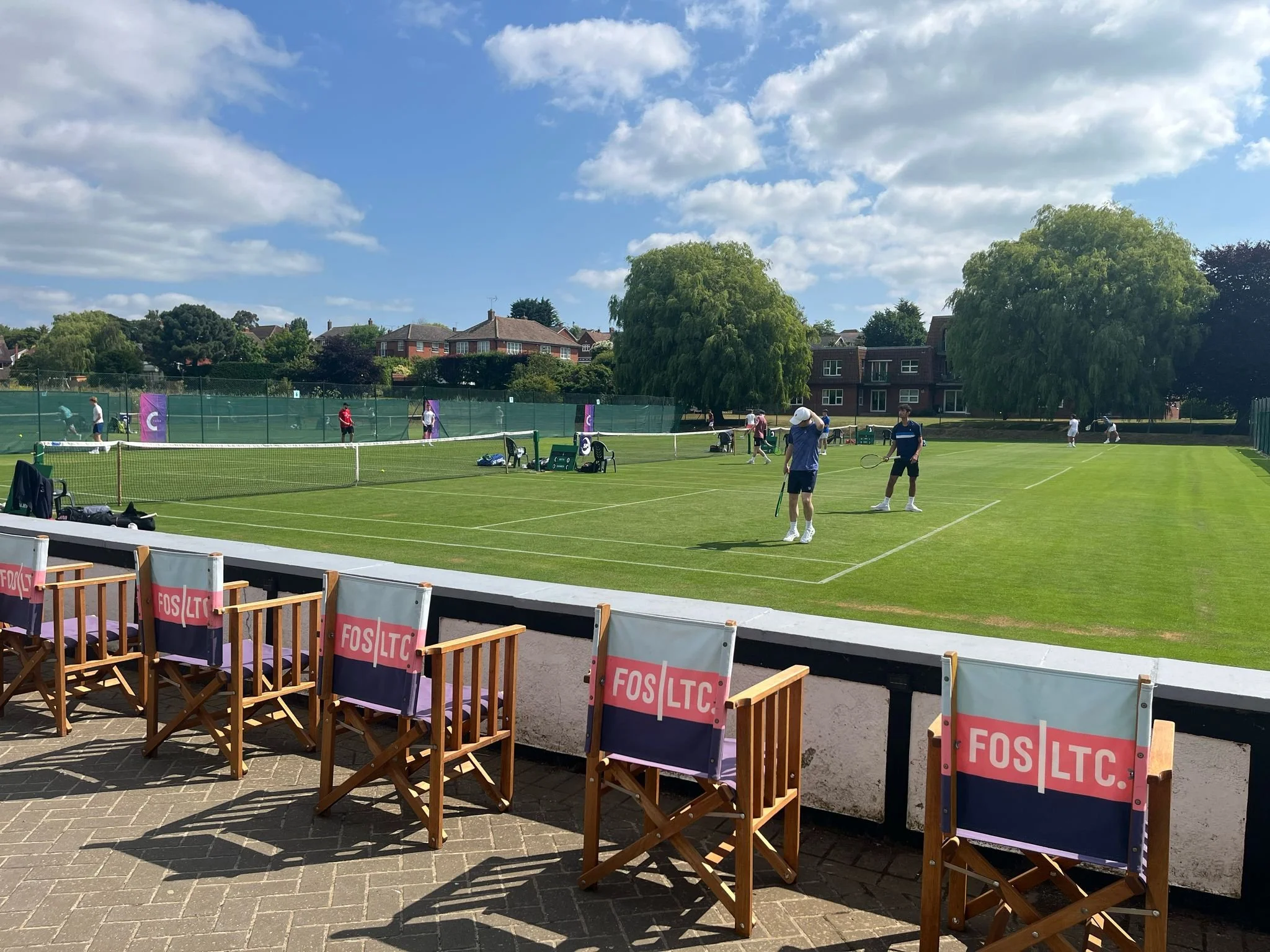 People playing tennis on an outdoor court on a sunny day with trees and houses in the background.