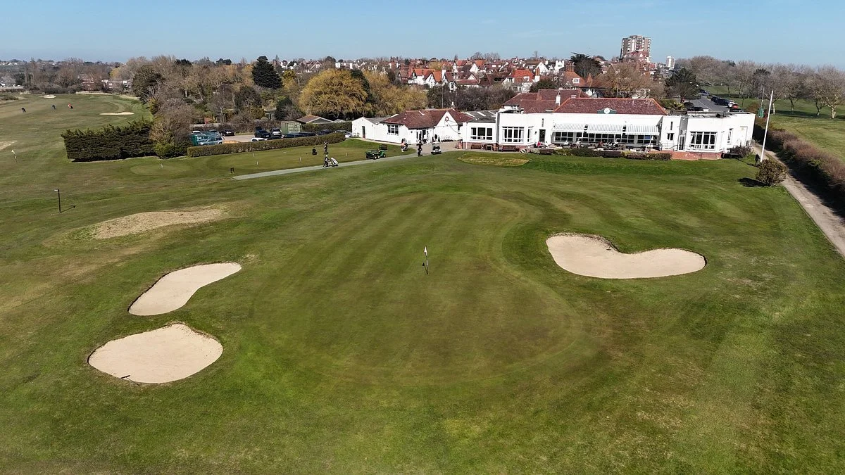 Aerial view of a golf course green with three sand bunkers, a flagstick, and a clubhouse building in the background, surrounded by trees and residential houses.