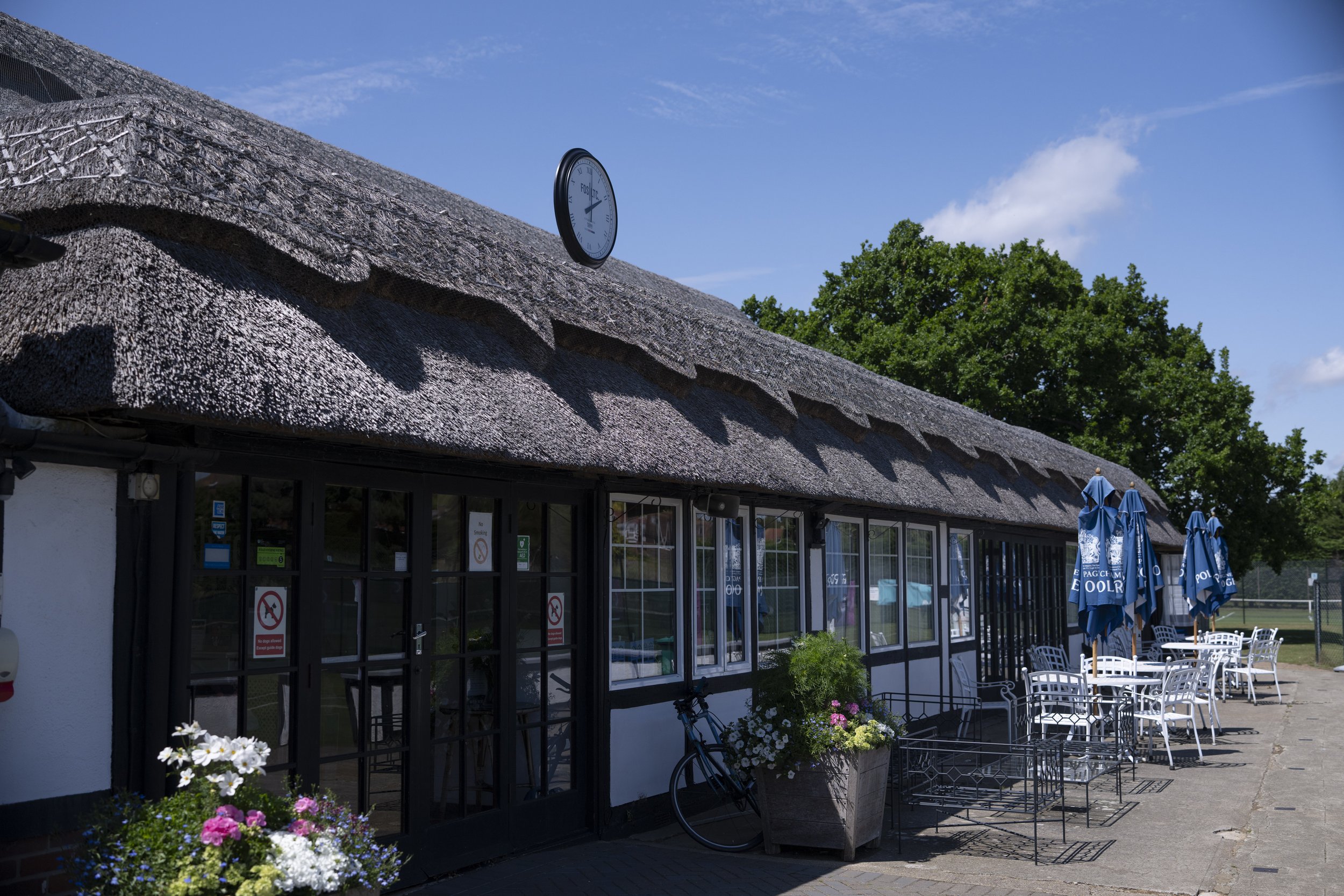 Exterior view of a building with a thatched roof, glass doors, and outdoor seating with tables, chairs, and umbrellas.