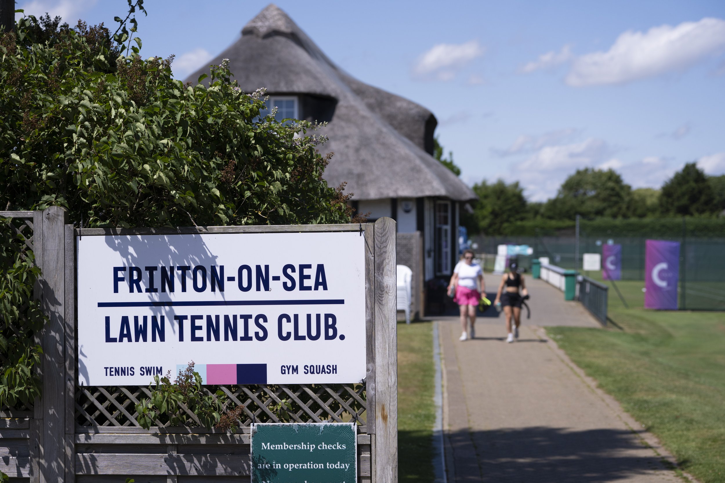 Sign for Frinton-on-Sea Lawn Tennis Club with people walking on a pathway in front of a thatched-roof building, tennis courts with purple banners, and a partly cloudy sky.
