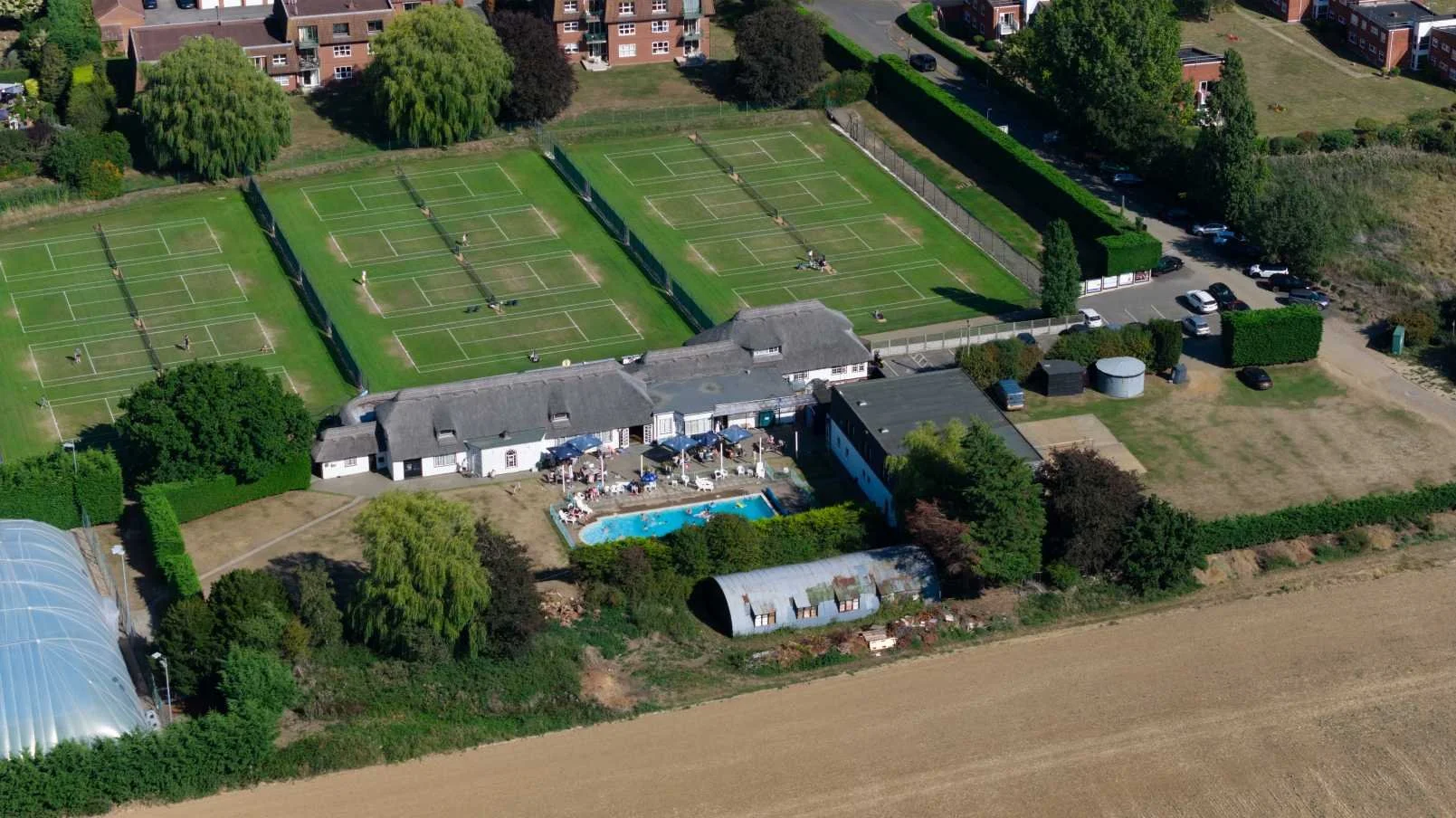 Aerial view of a facility with tennis courts, a swimming pool, a greenhouse, and surrounding greenery and residential buildings.