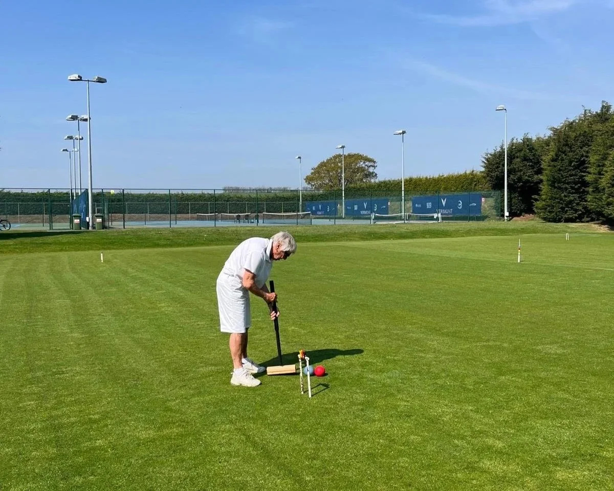 An elderly man dressed in white shorts and shirt playing lawn croquet on a grassy field on a sunny day.