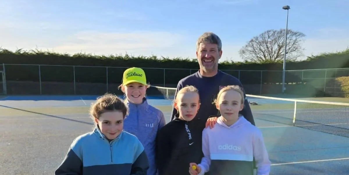 A man and four young girls standing on a tennis court outdoors, smiling at the camera. The man is in the back center, and the girls are in front of him. One girl is wearing a yellow Titleist cap, and another has a black hoodie. The background shows a blue sky, trees, and tennis net.