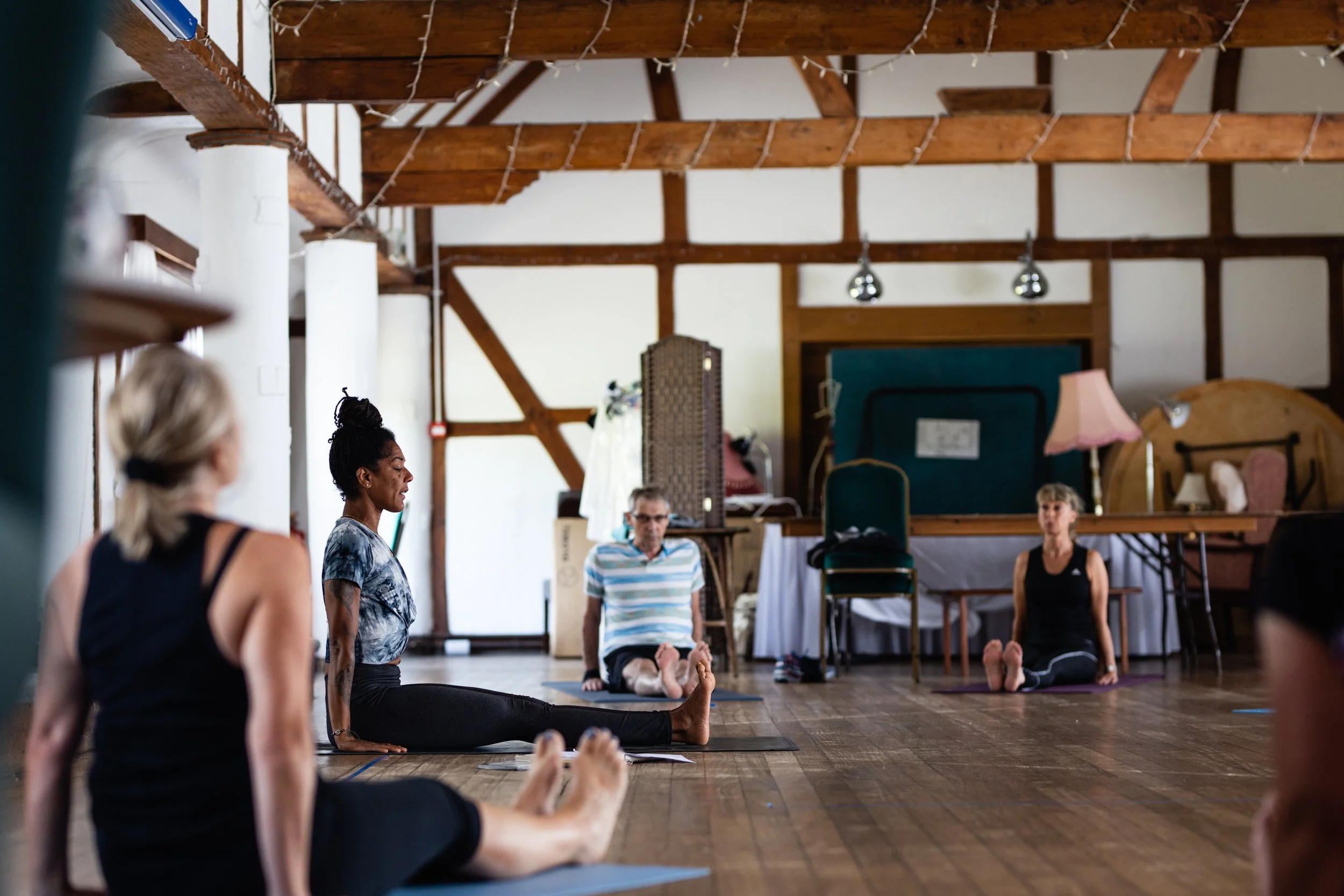 A group of people practicing yoga indoors in a studio with wooden beams on the ceiling. They are sitting on yoga mats in a seated forward bend pose.