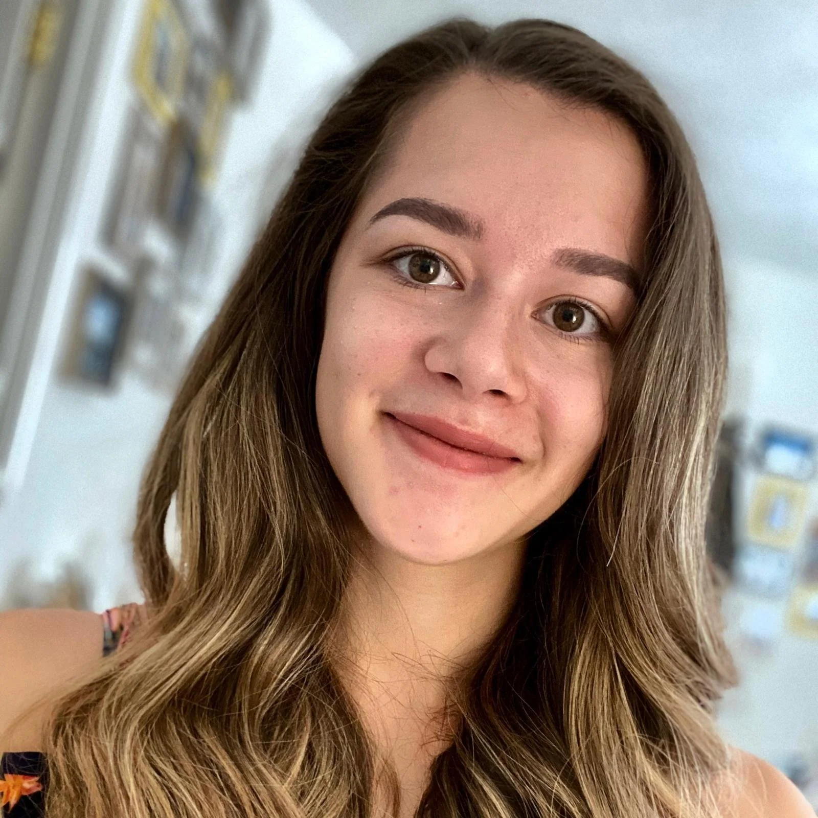 A young woman with long, wavy brown hair and brown eyes, smiling indoors with a blurred background of frames and decorations.