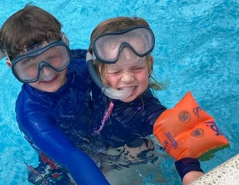 Two children in a swimming pool wearing goggles and swim gear, smiling at the camera.