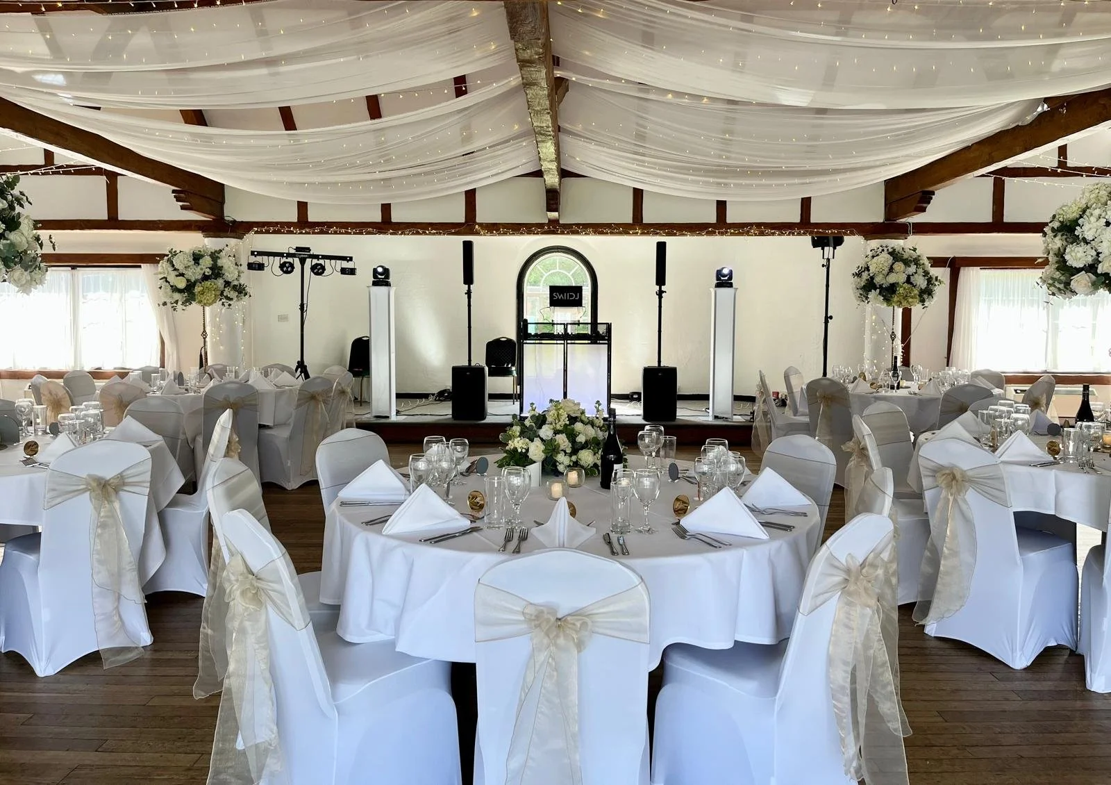 A decorated banquet hall with round tables covered with white tablecloths and chairs adorned with white fabric and bows. The centerpieces feature white flowers. There is a stage at the front with musical equipment and lighting, and draped fabric and string lights decorate the ceiling.