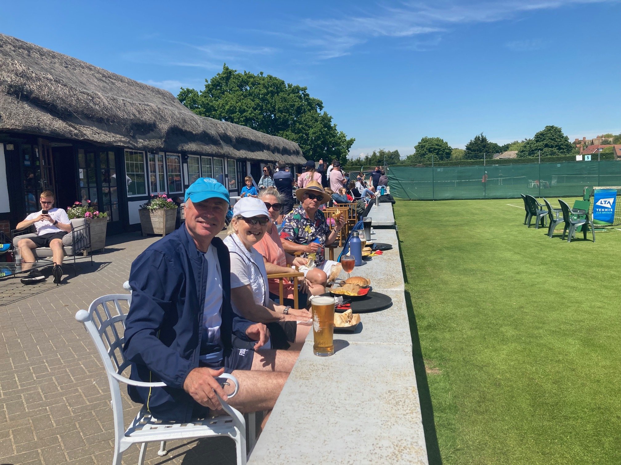 People sitting at a long table outdoors at a tennis club enjoying drinks and food on a sunny day, with tennis courts and a thatched-roof building in the background.