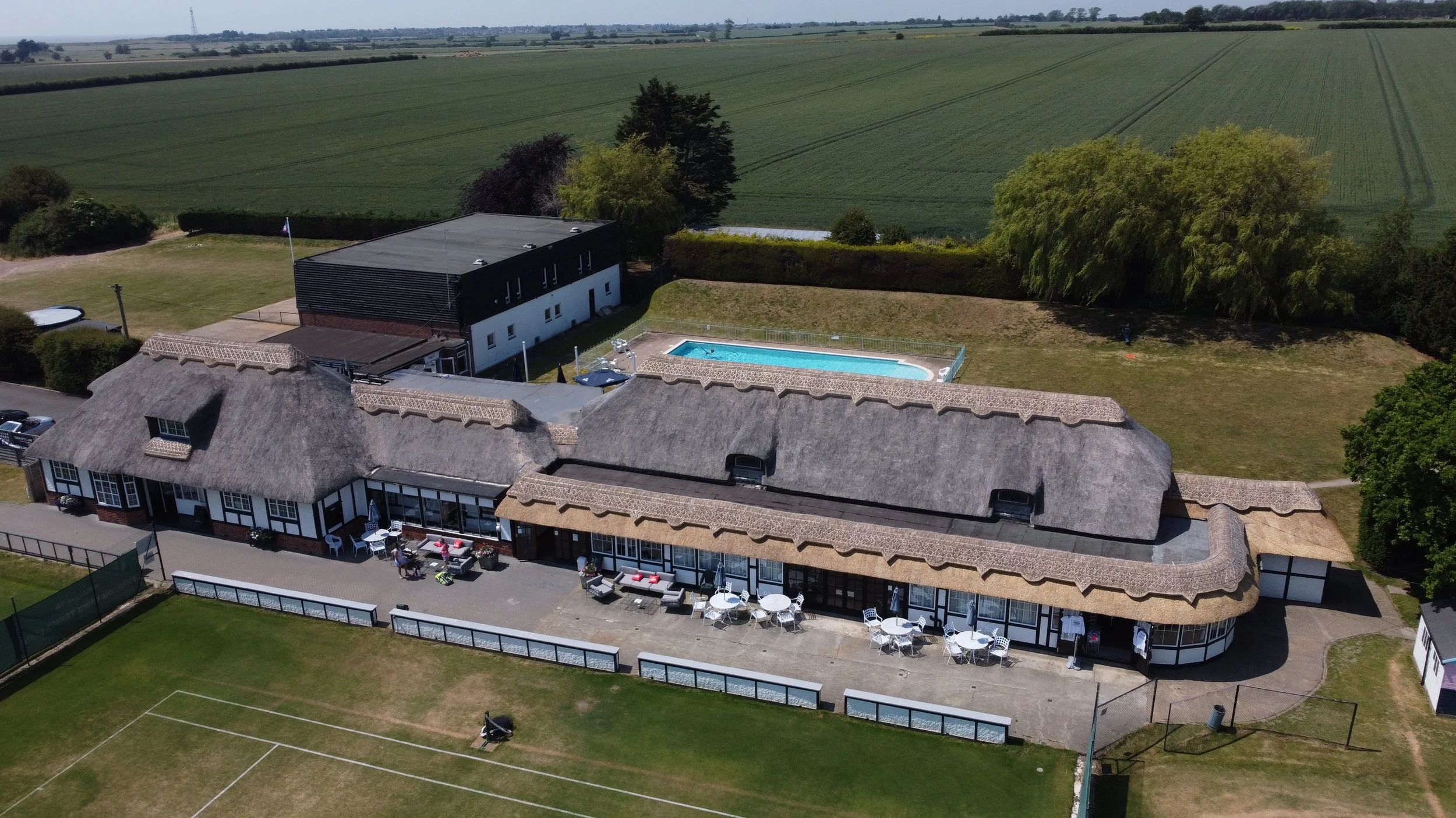 Aerial view of a large house with a thatched roof, outdoor patio with tables and umbrellas, swimming pool, tennis court, and surrounding greenery and fields.