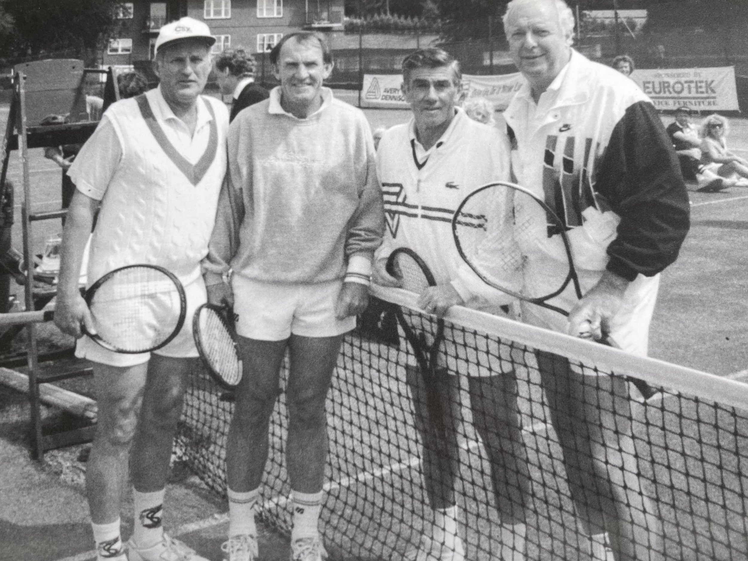 Four men standing on a tennis court posed for a photo, holding tennis rackets; two of them are dressed in tennis attire, and the other two are in casual clothes. There is a net in front of them, and a couple of people seated in the background.