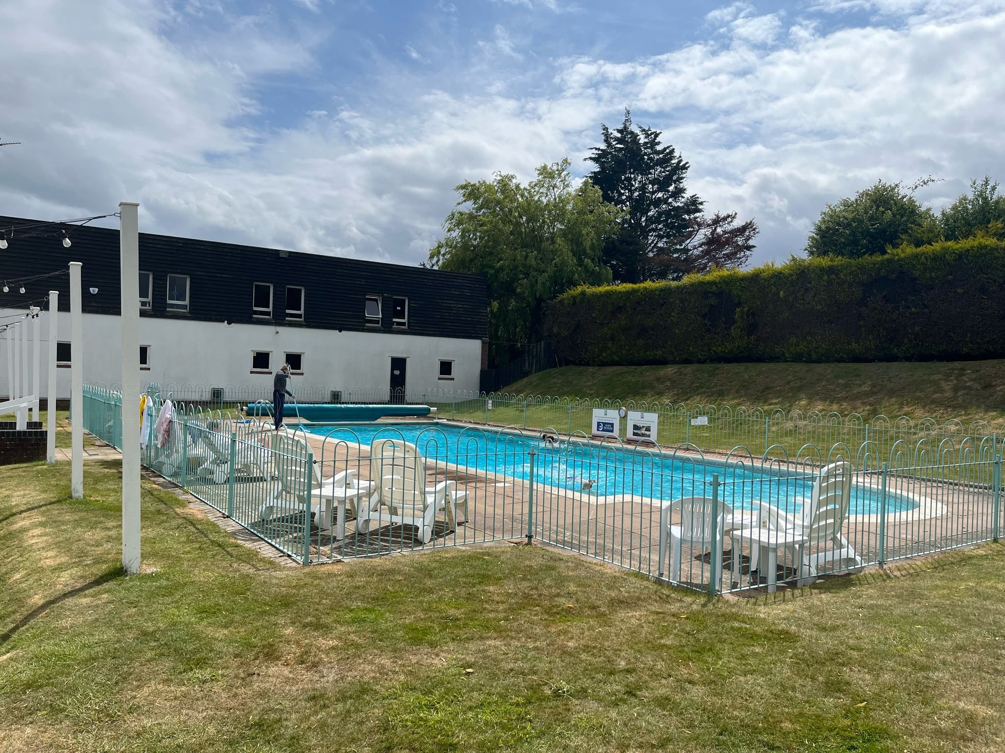An outdoor swimming pool fenced with a white metal fence, with plastic lounge chairs and tables nearby. A person is cleaning the pool with a long pole, and there is a black building with small windows in the background, with trees and a cloudy sky above.