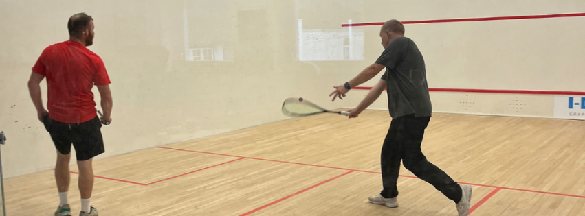 Two people playing squash in an indoor court, one preparing to hit the ball with a racket while the other stands nearby watching.