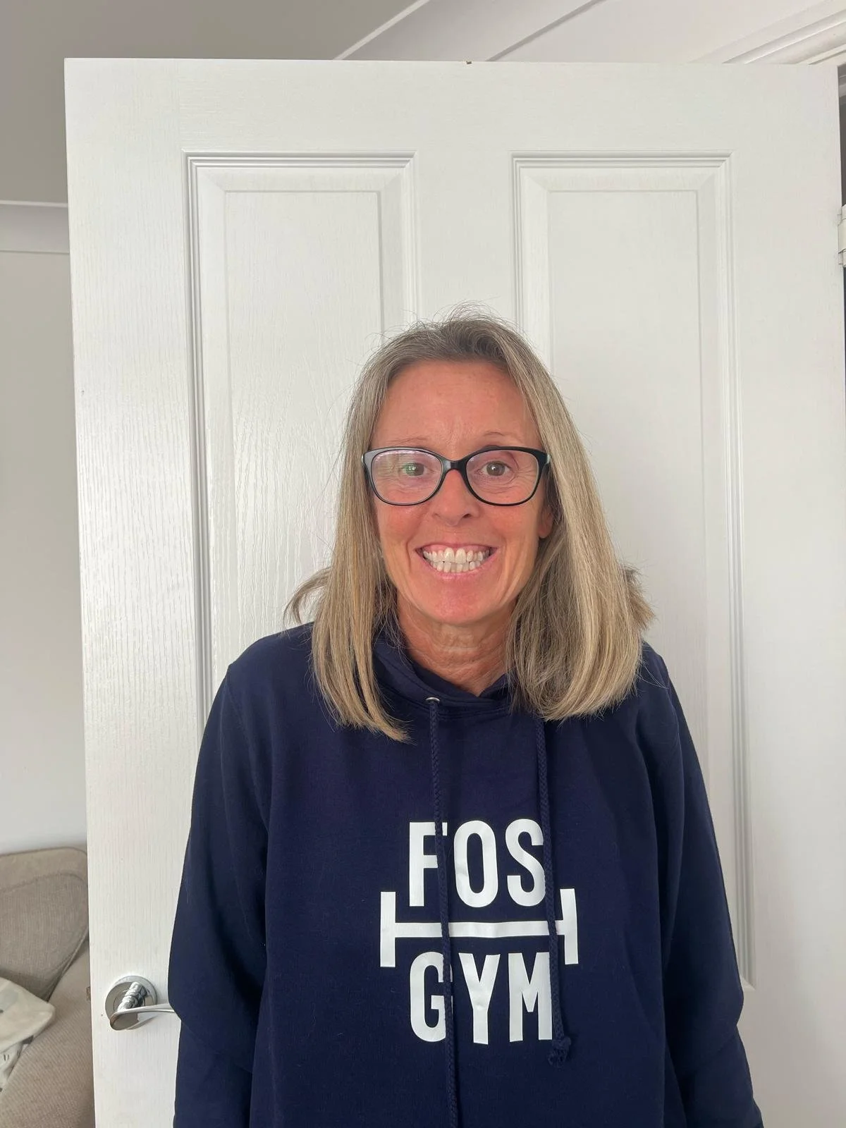 Smiling woman with glasses wearing a navy blue hoodie that says 'FOS GYM' in white letters, standing in front of a white door.