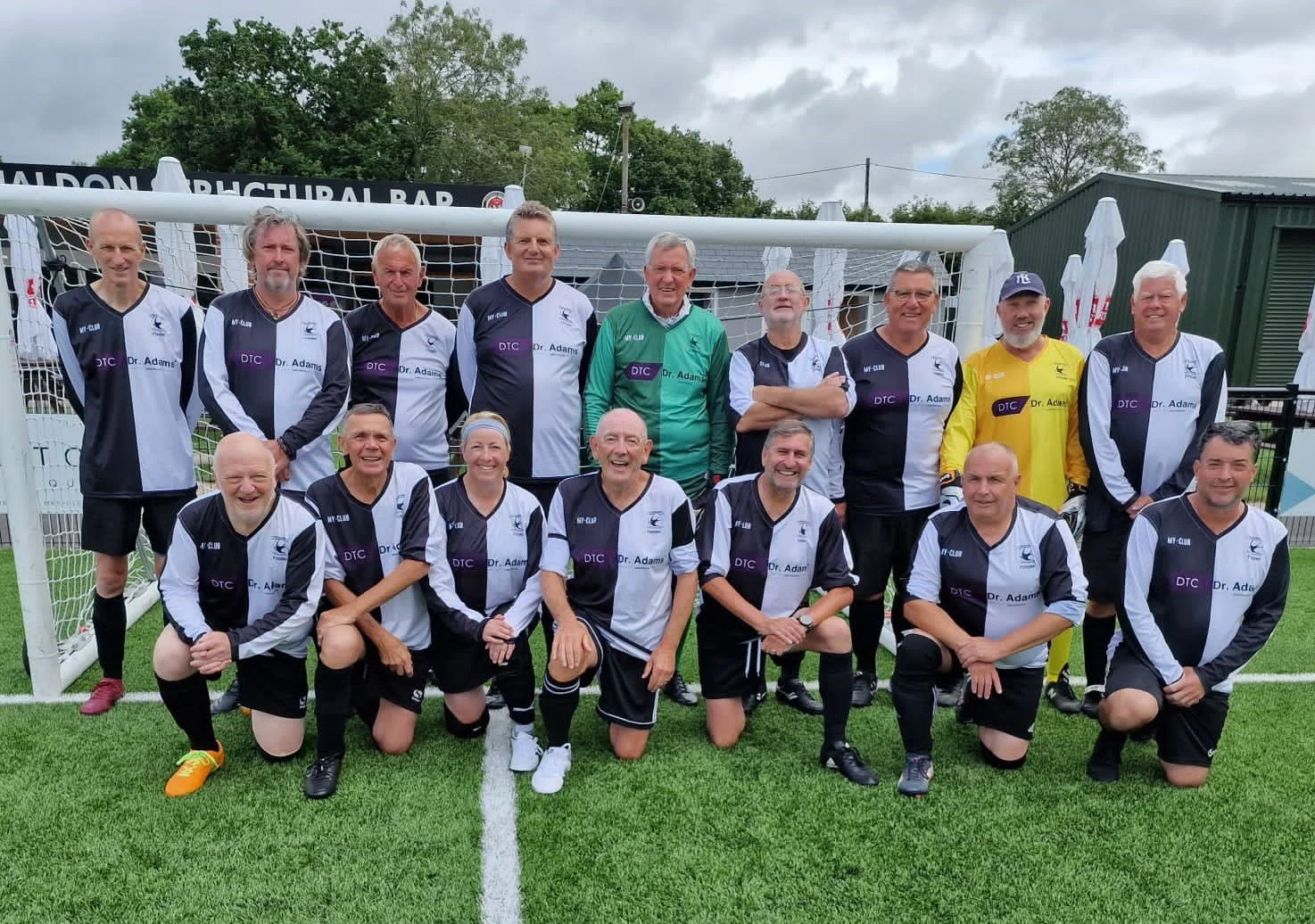 Group of adult men and women in soccer jerseys on a green field, posing for a team photo in front of a goal.
