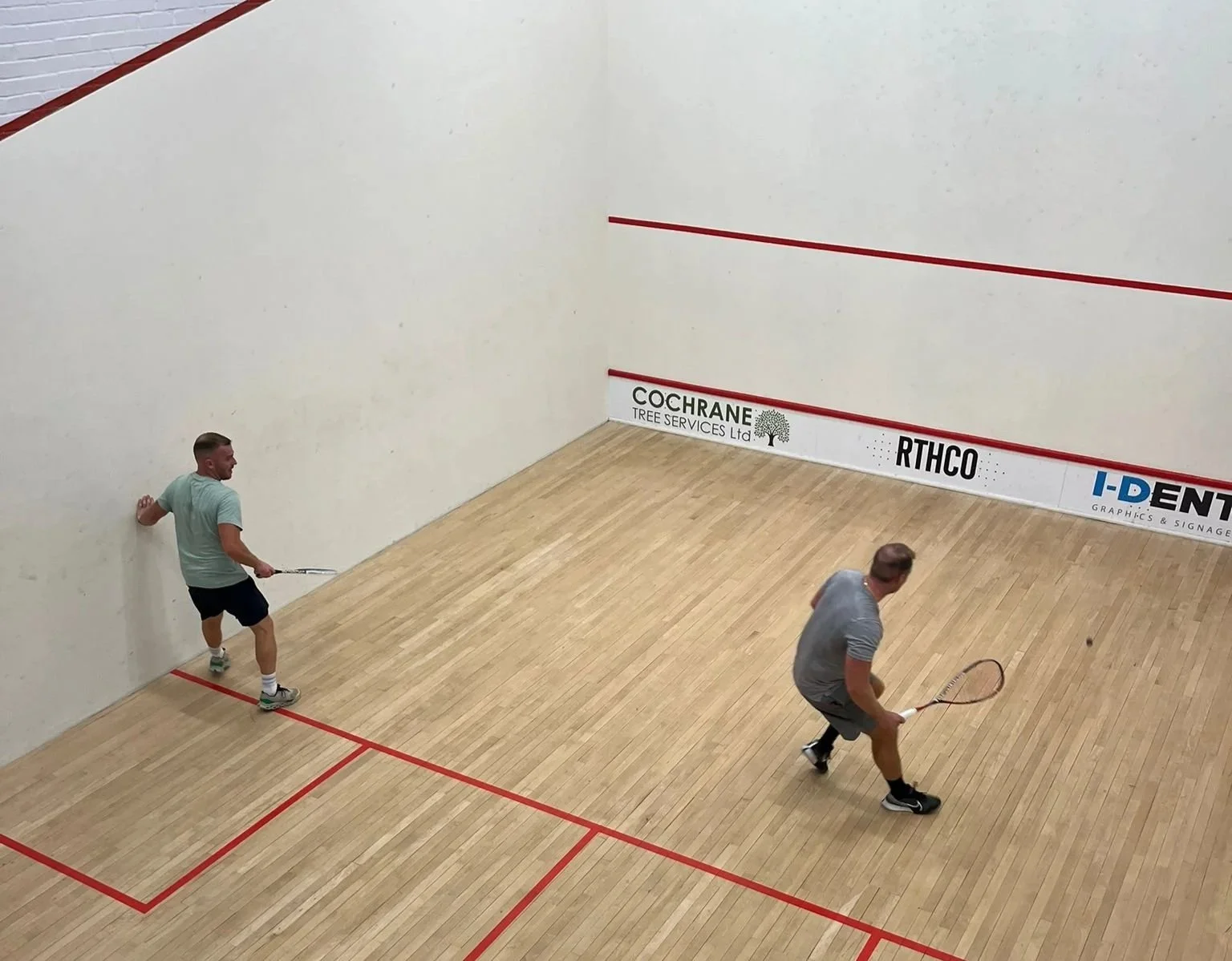 Two men playing squash on an indoor court with wooden floors and white walls, markings in red, and advertising banners at the side.