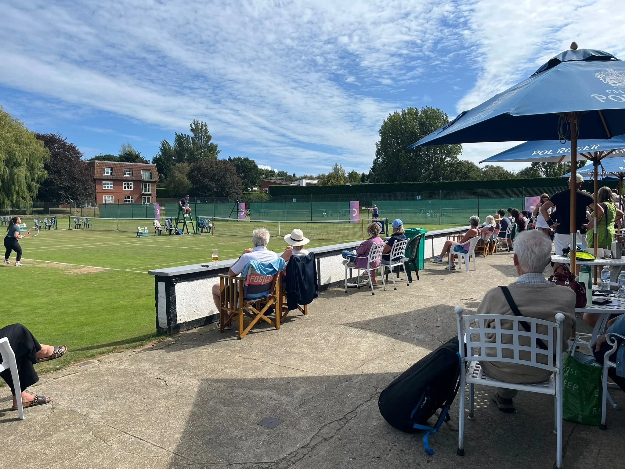 People watching a tennis match from chairs and tables outside on a sunny day with umbrellas, tennis courts, a blue sky, and trees in the background.