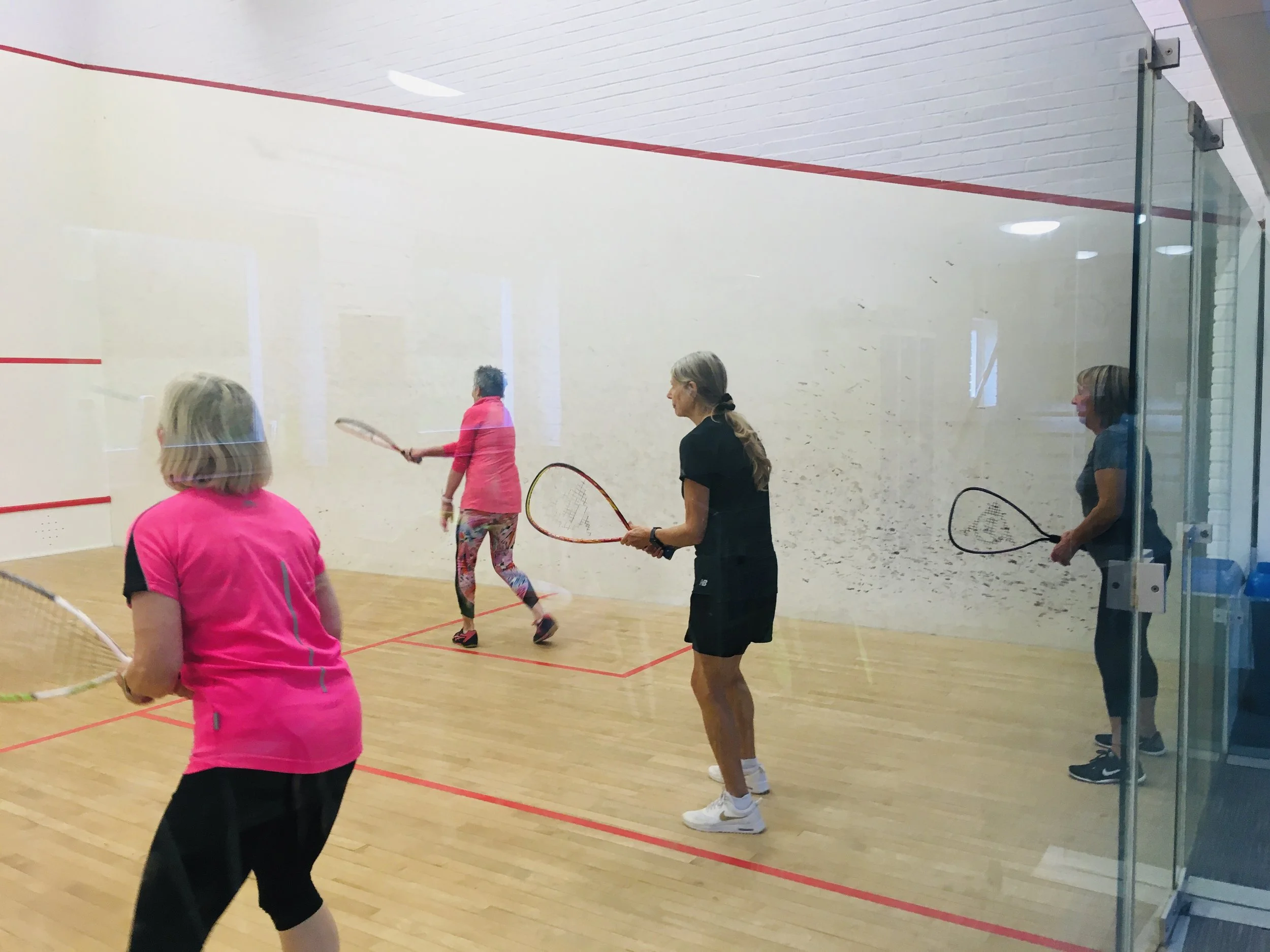 Four women playing squash on an indoor court with a glass wall, with two women near the front and two at the back, holding squash rackets, in athletic clothing.