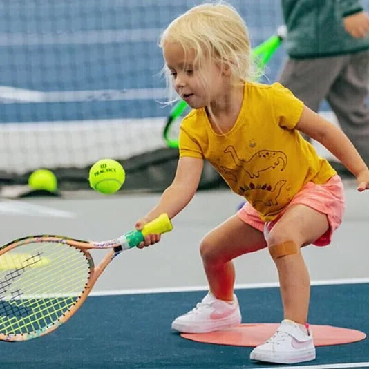 A young girl in a yellow t-shirt and pink shorts playing tennis on an indoor court, holding a tennis racket and reaching for a yellow tennis ball.